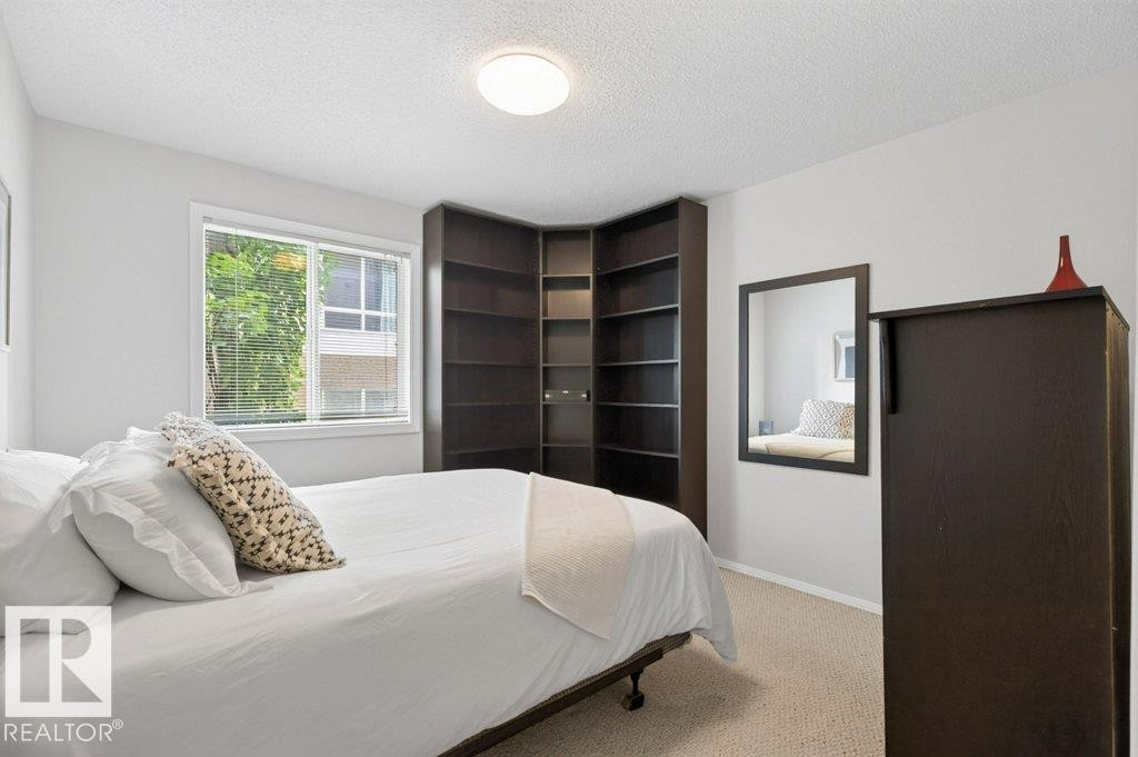 Window with blinds, wall-mounted mirror, built-in corner shelving, overhead flush-mount lighting, and wall-to-wall carpeting - #201 10528 77 Av Nw, Edmonton, AB - Indoor Photo Showing Bedroom