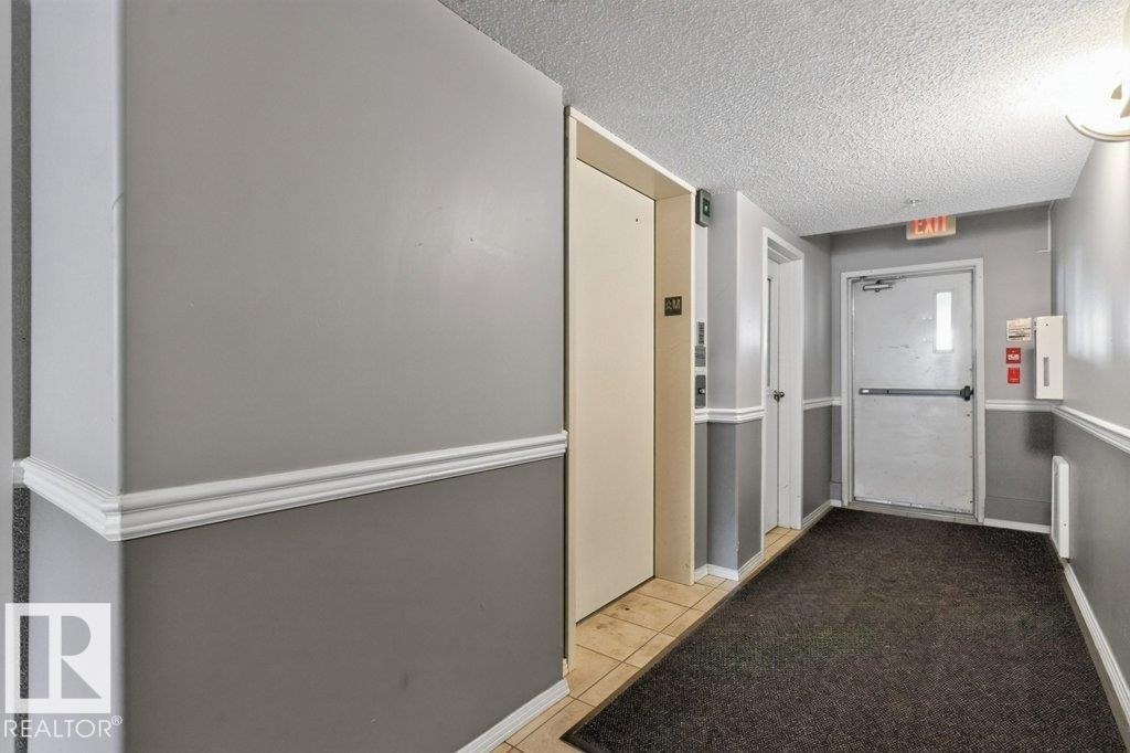 Well-Maintained Common Hallway with Neutral Palette and Practical Layout This institutional corridor features clean white and gray two-tone wall treatment with a protective chair rail, dark carpet flooring, and wood-finish door frames - #201 10528 77 Av Nw, Edmonton, AB - Indoor Photo Showing Other Room