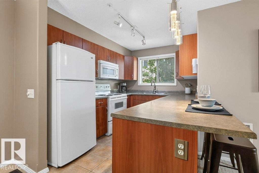 Kitchen featuring wood-finish cabinetry, white appliances, track lighting, and a peninsula with a stone-look countertop - #201 10528 77 Av Nw, Edmonton, AB - Indoor Photo Showing Kitchen