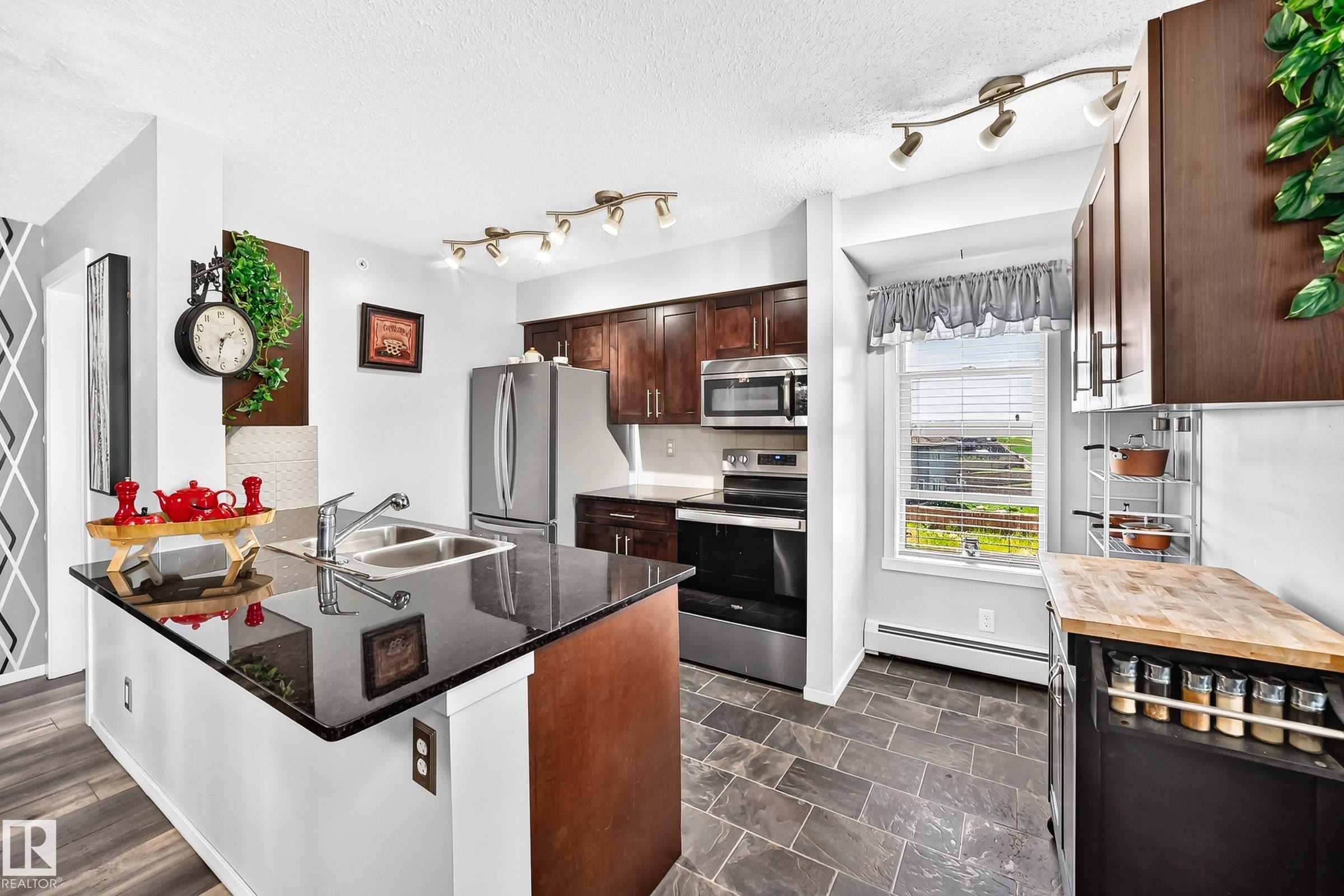 Kitchen featuring dark wood-finish cabinetry, stainless steel appliances, black countertops, a dual basin sink, and slate-style floor tiling - #401 5390 Chappelle Rd Sw, Edmonton, AB