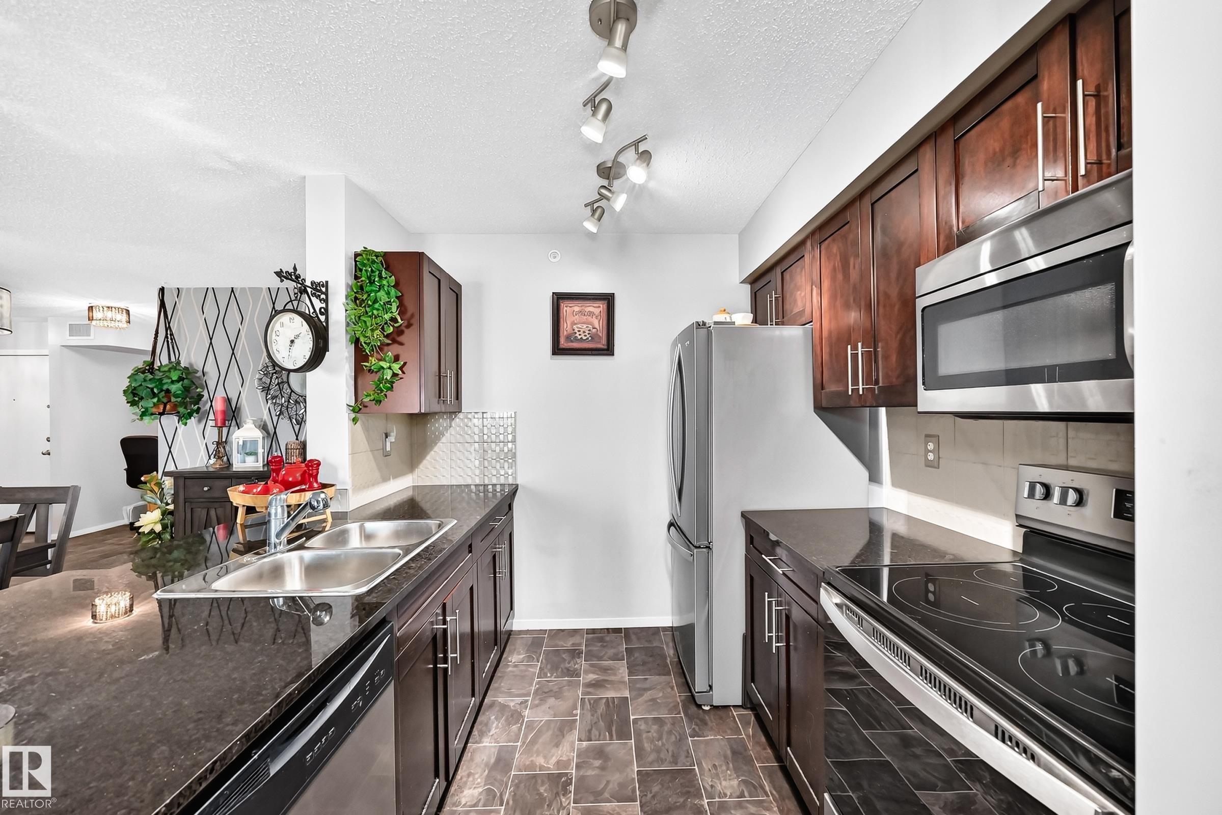 Kitchen featuring dark wood-finish cabinetry, stainless steel appliances, dark stone countertops, and tile flooring - #401 5390 Chappelle Rd Sw, Edmonton, AB