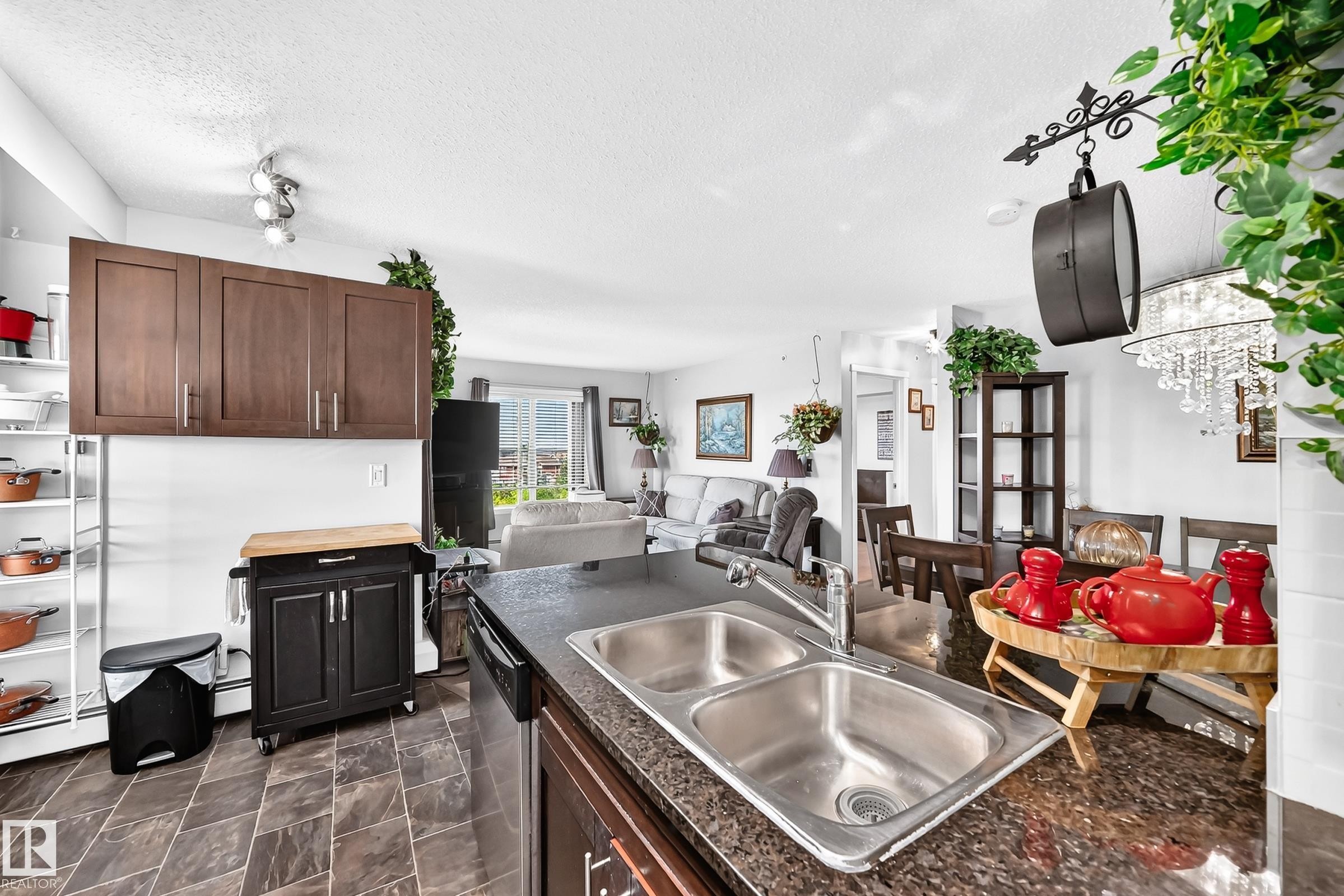 Open-concept kitchen with dark cabinetry, granite-style countertops, and a double stainless steel sink - #401 5390 Chappelle Rd Sw, Edmonton, AB
