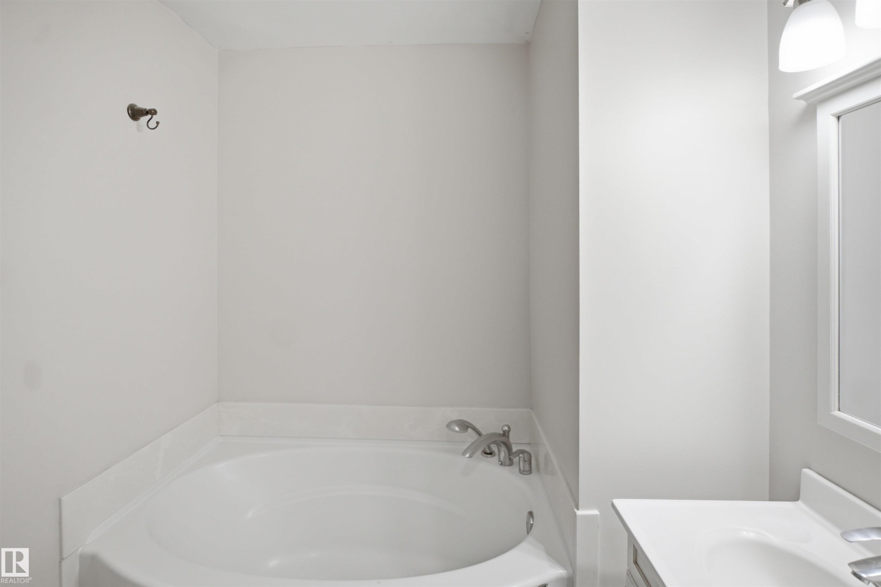 Corner soaking tub with polished chrome fixtures and an adjacent vanity featuring a rectangular sink, white countertop, and a framed mirror with overhead lighting - 3515 20 Ave Nw, Edmonton, AB