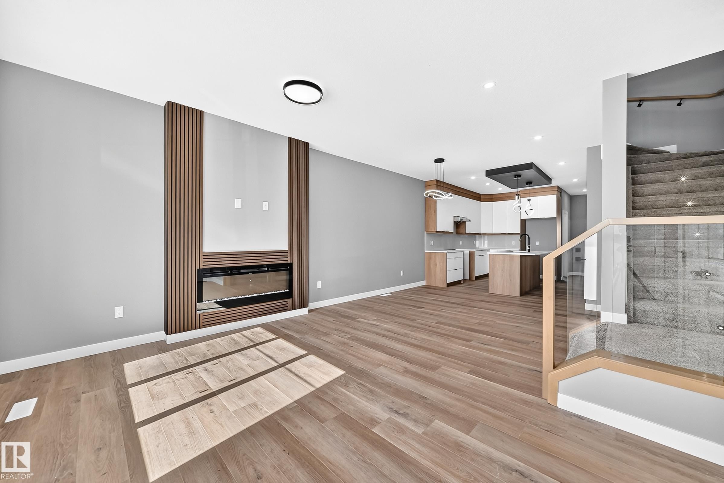 Wood-finish flooring throughout, contemporary fireplace with wood slat accent wall, modern kitchen with two-tone cabinetry and island, and a staircase featuring glass panel railings - 426 Crystal Creek Link, Leduc, AB - Indoor With Fireplace