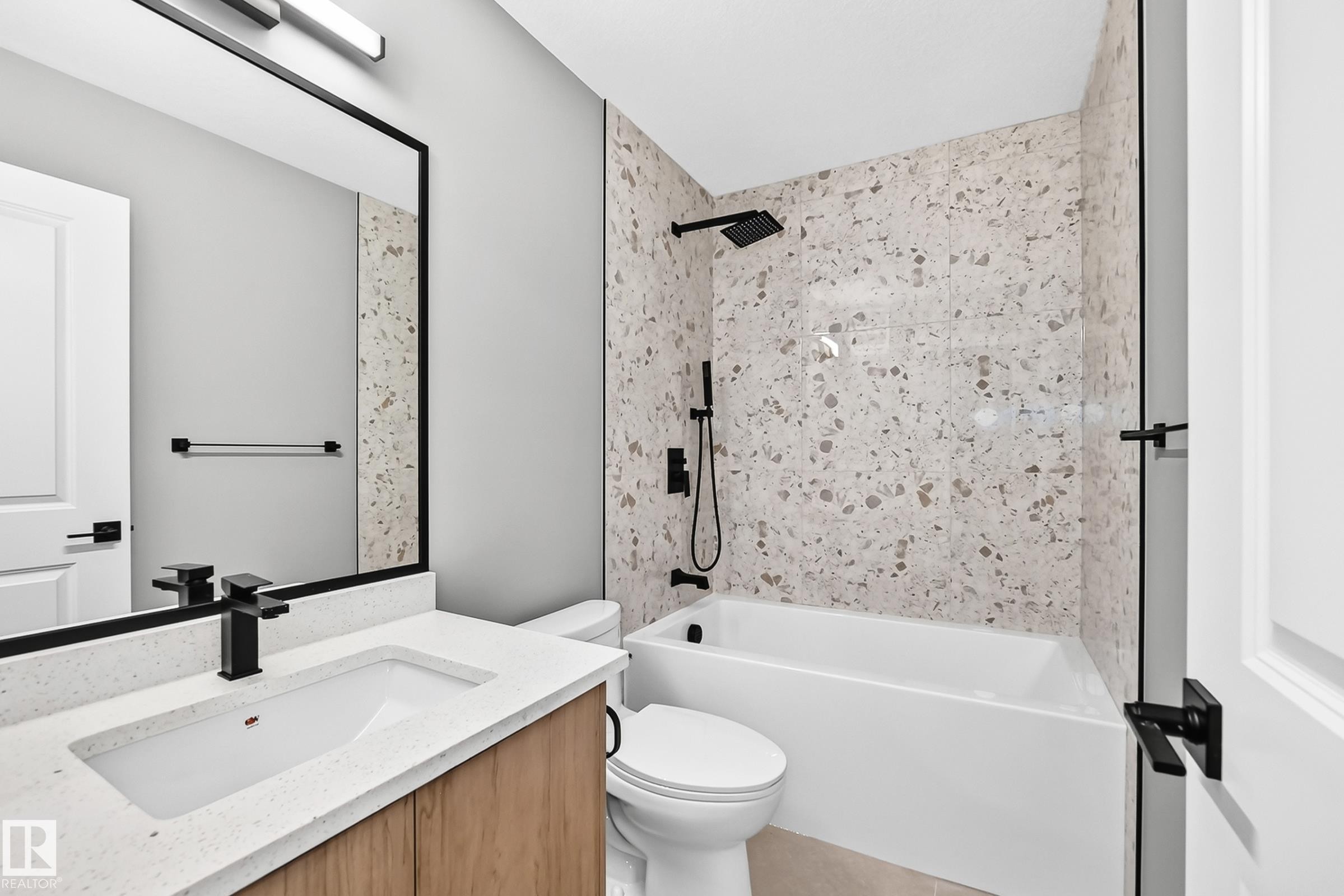 Bathroom featuring a single-sink vanity with a light-toned countertop, a rectangular mirror with a black frame, a bathtub with a tiled surround, a toilet, and black fixtures - 426 Crystal Creek Link, Leduc, AB - Indoor Photo Showing Bathroom