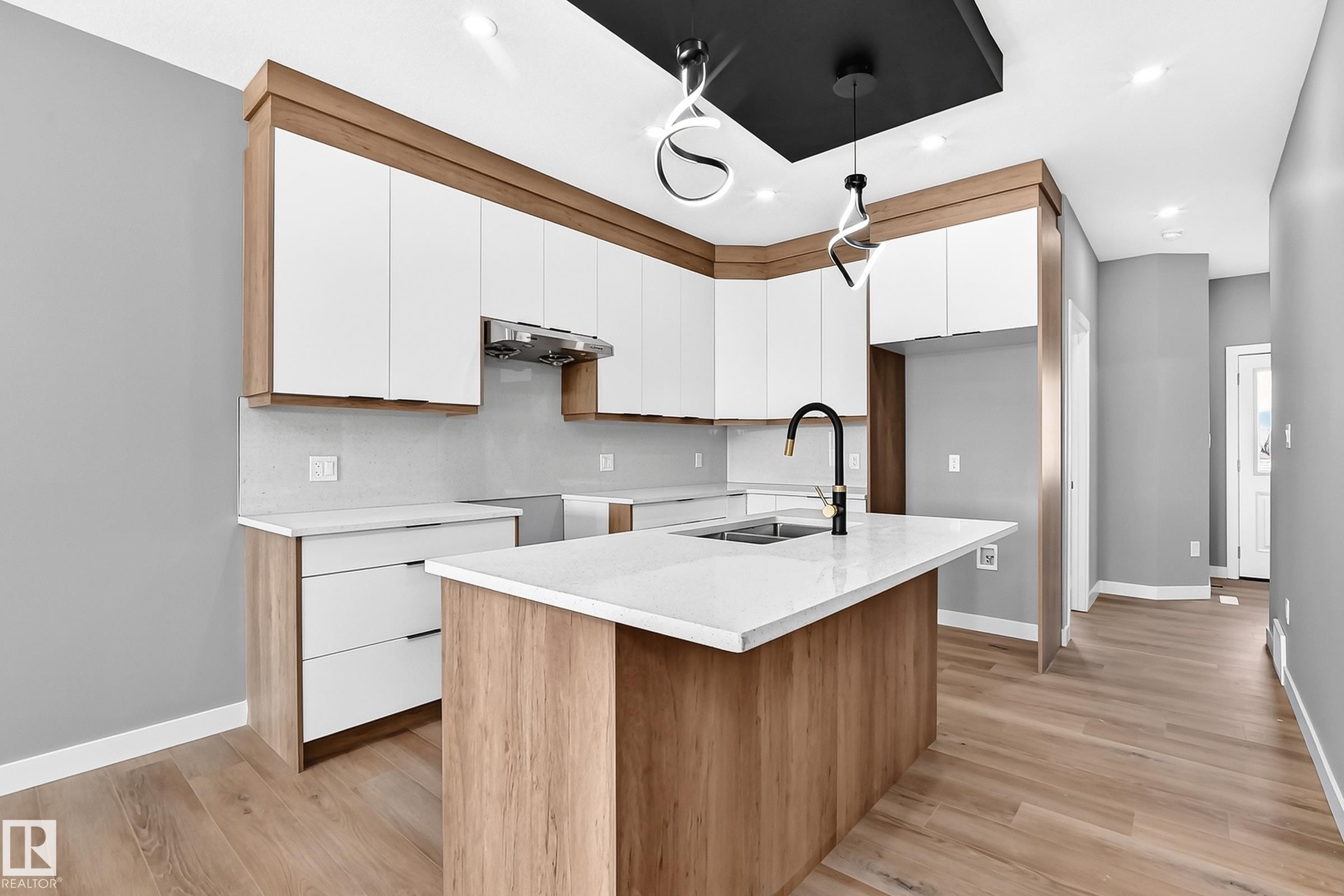 Kitchen featuring wood-finish flooring, white cabinetry with wood accents, a white countertop island with an integrated sink, a contemporary black faucet, and pendant lighting fixtures - 426 Crystal Creek Link, Leduc, AB - Indoor Photo Showing Kitchen With Double Sink
