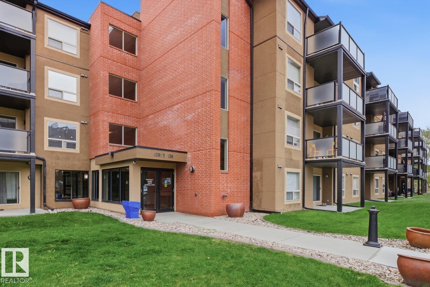Multi-story building featuring a brick and stucco facade, glass-paneled balconies, and a concrete walkway - #106 13907 136 St Nw, Edmonton, AB - Outdoor With Exterior