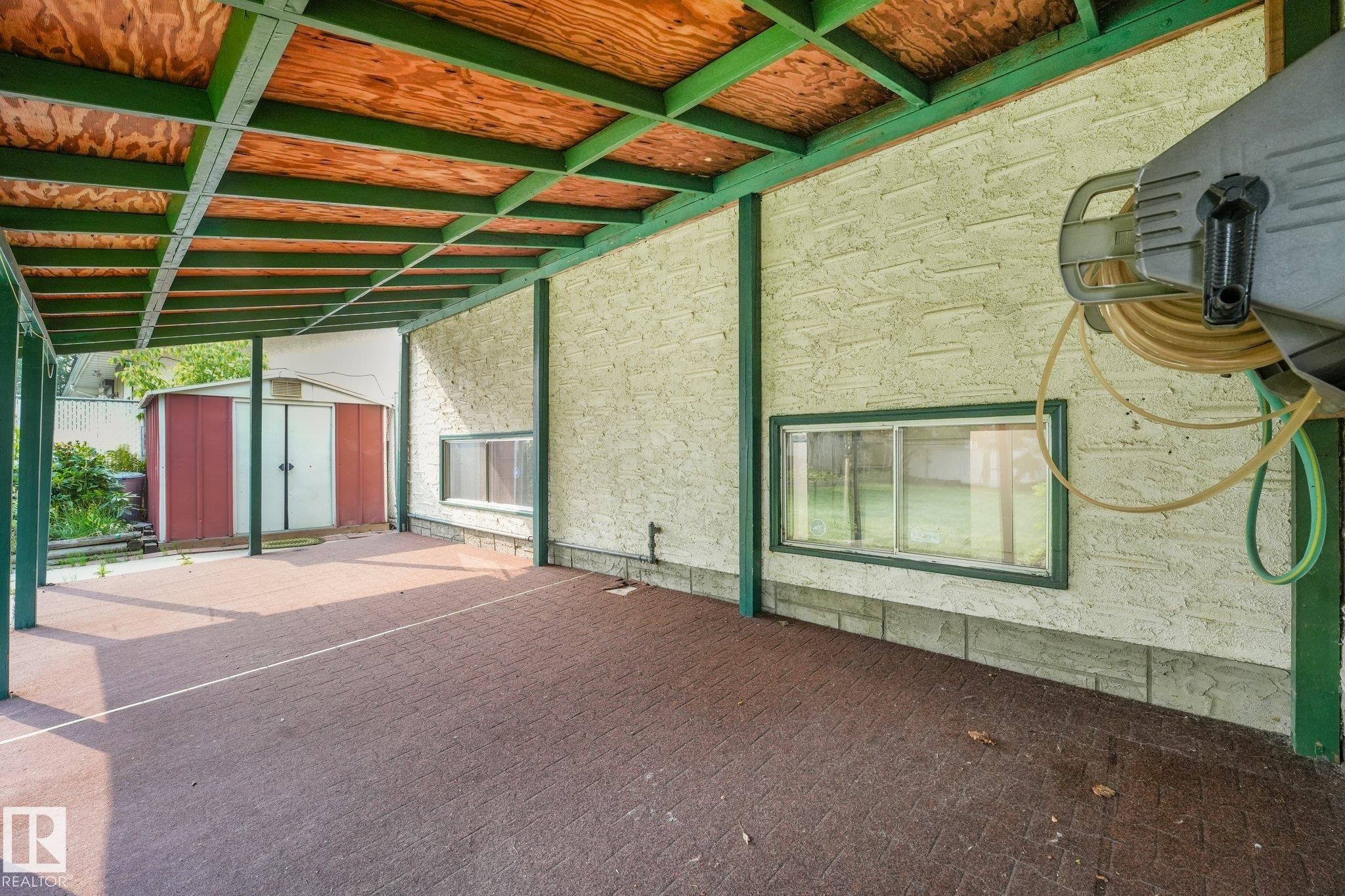 Covered patio featuring an exposed plywood ceiling with green structural beams, a textured exterior wall, and a patterned paver floor - 4135 134 Avenue Nw, Edmonton, AB - Outdoor With Exterior