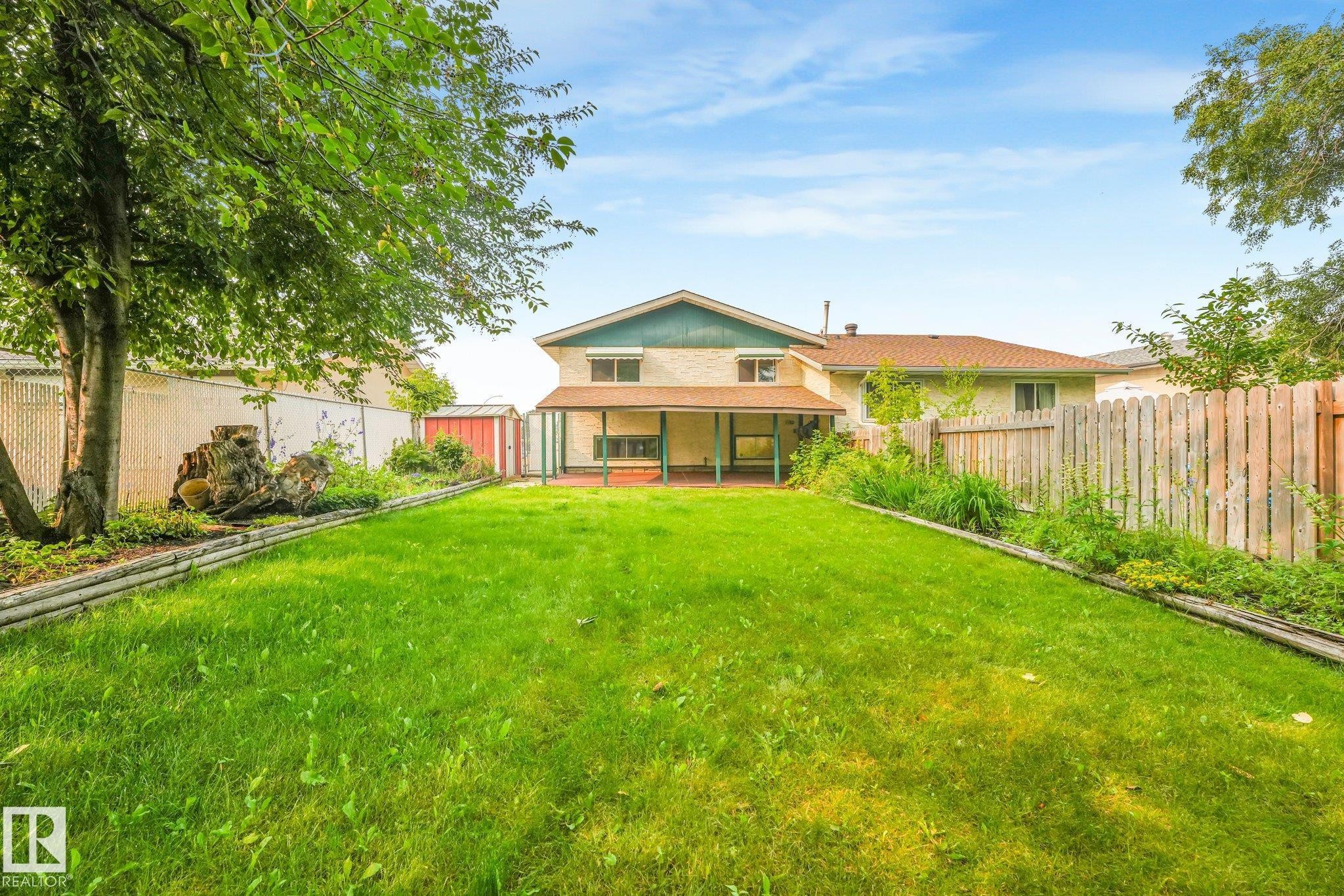 Expansive grassy backyard featuring a covered patio, wood fencing, mature trees, and a storage shed - 4135 134 Avenue Nw, Edmonton, AB - Outdoor With Deck Patio Veranda