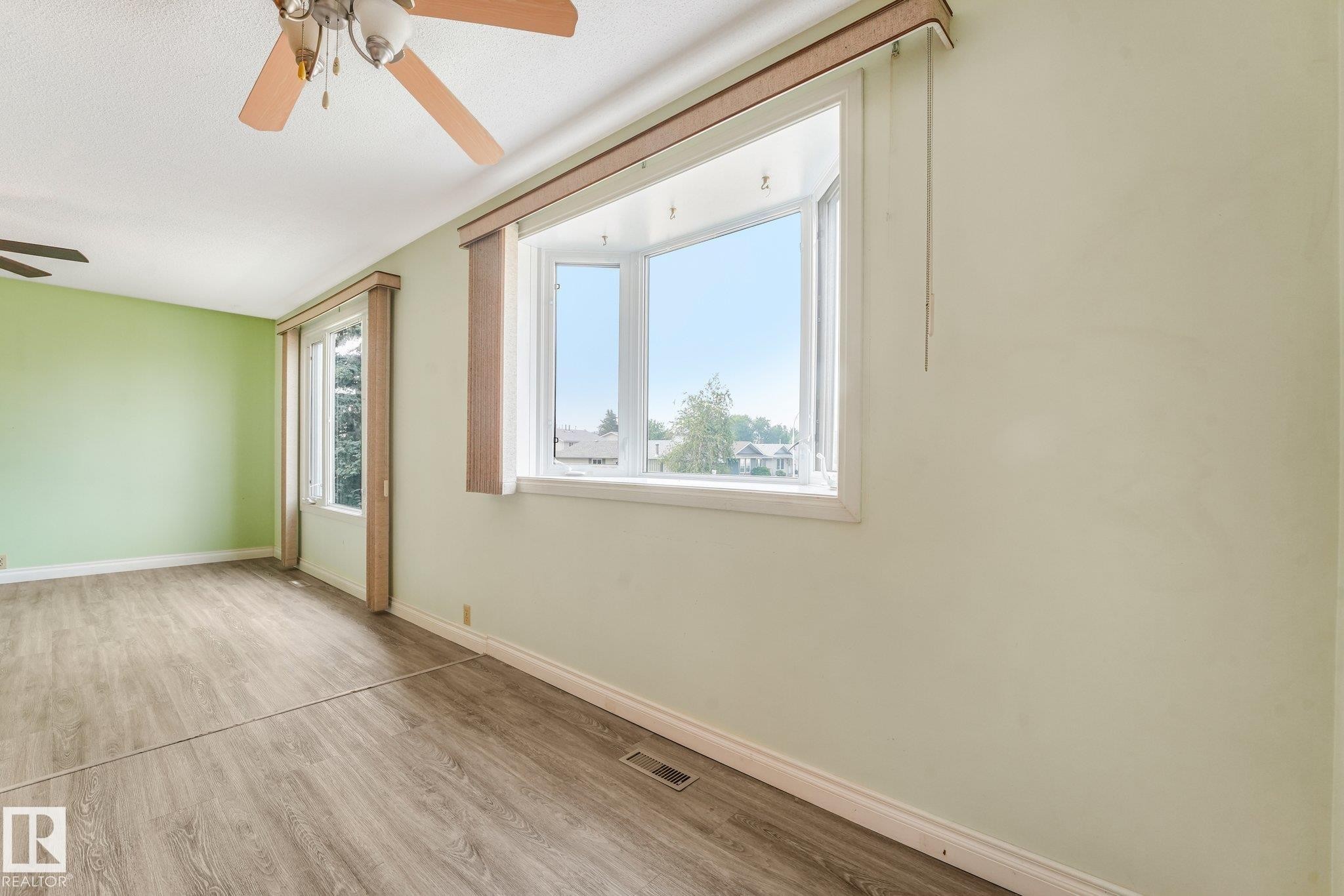 Spacious room featuring a bay window, two standard windows, wood-finish flooring, light green wall paint, and a ceiling fan - 4135 134 Avenue Nw, Edmonton, AB - Indoor Photo Showing Other Room
