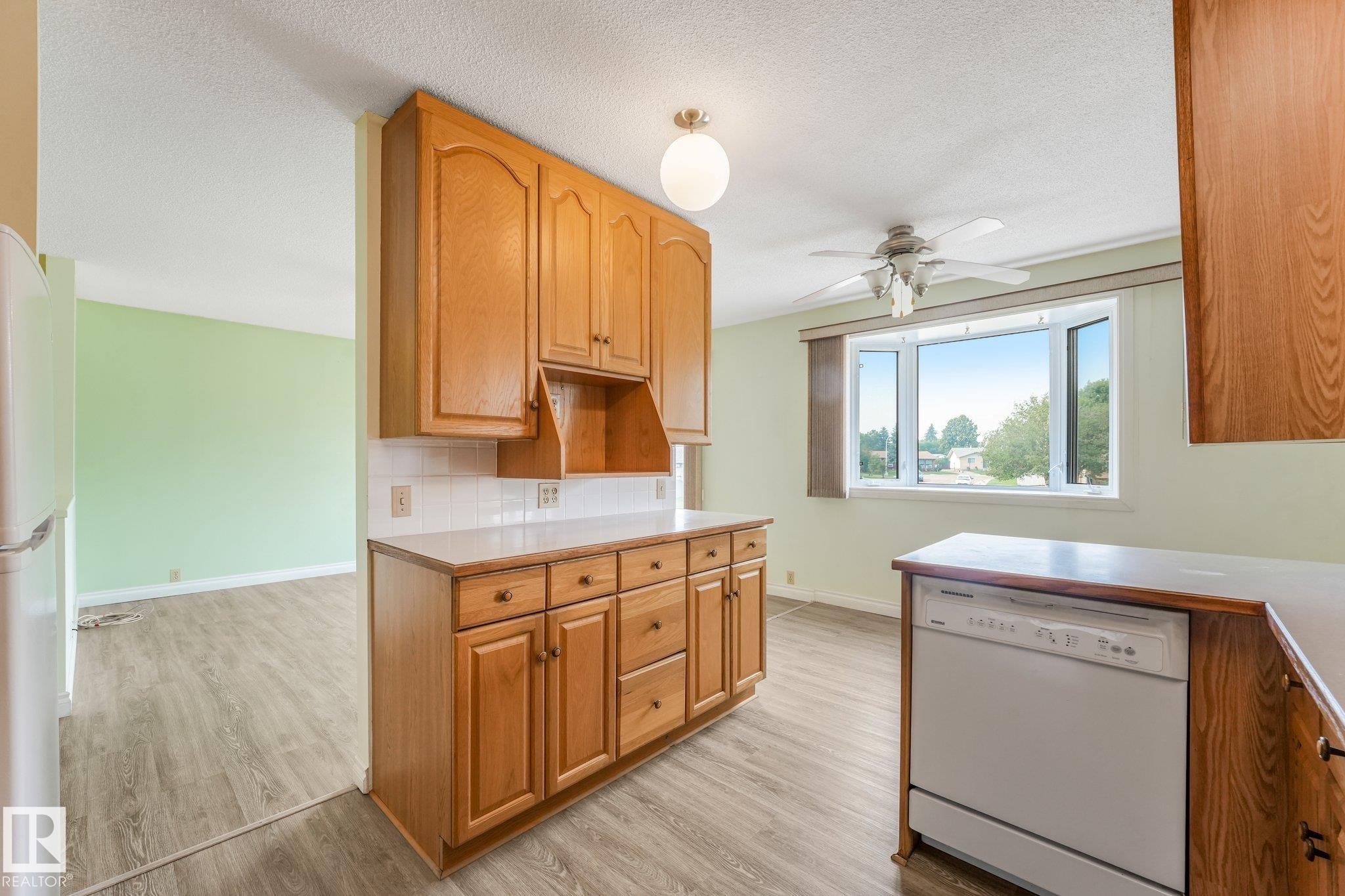 Kitchen featuring wood cabinetry, white tile backsplash, light-toned countertops, and wood-finish flooring - 4135 134 Avenue Nw, Edmonton, AB - Indoor Photo Showing Kitchen