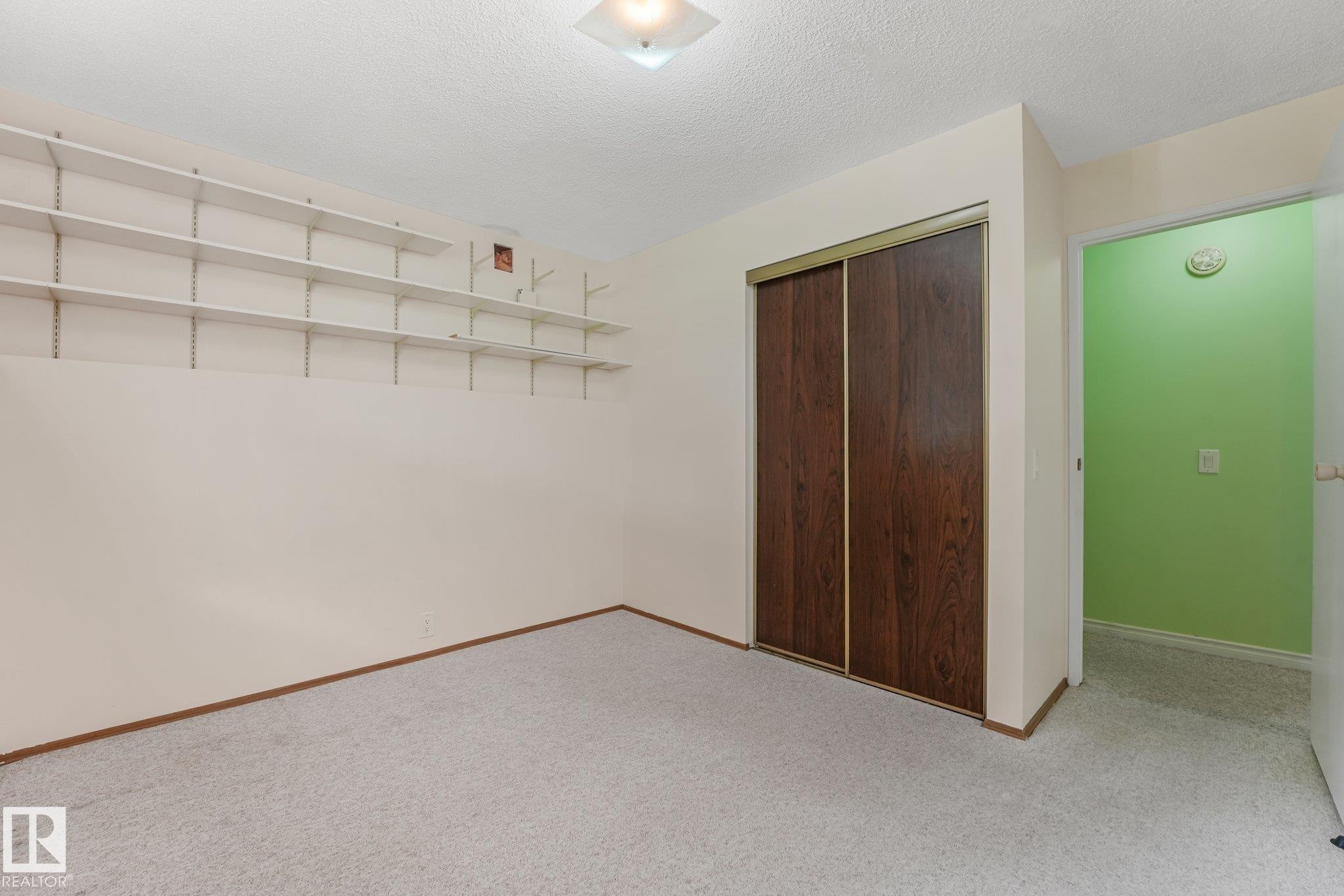Carpeted room featuring a ceiling-mounted light fixture, extensive wall-mounted shelving, wood-paneled sliding closet doors, and light-toned walls - 4135 134 Avenue Nw, Edmonton, AB - Indoor Photo Showing Other Room
