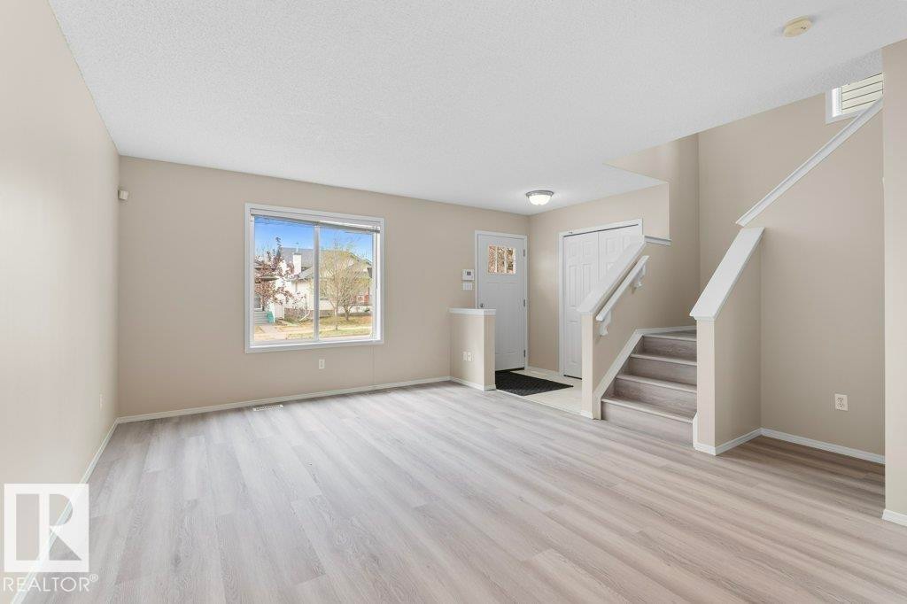 Expansive living area featuring light wood-finish flooring, a large window, neutral wall tones, a front entry door with decorative glass, and a staircase with white risers and handrails - 21344 88 Avenue Nw, Edmonton, AB - Indoor Photo Showing Other Room