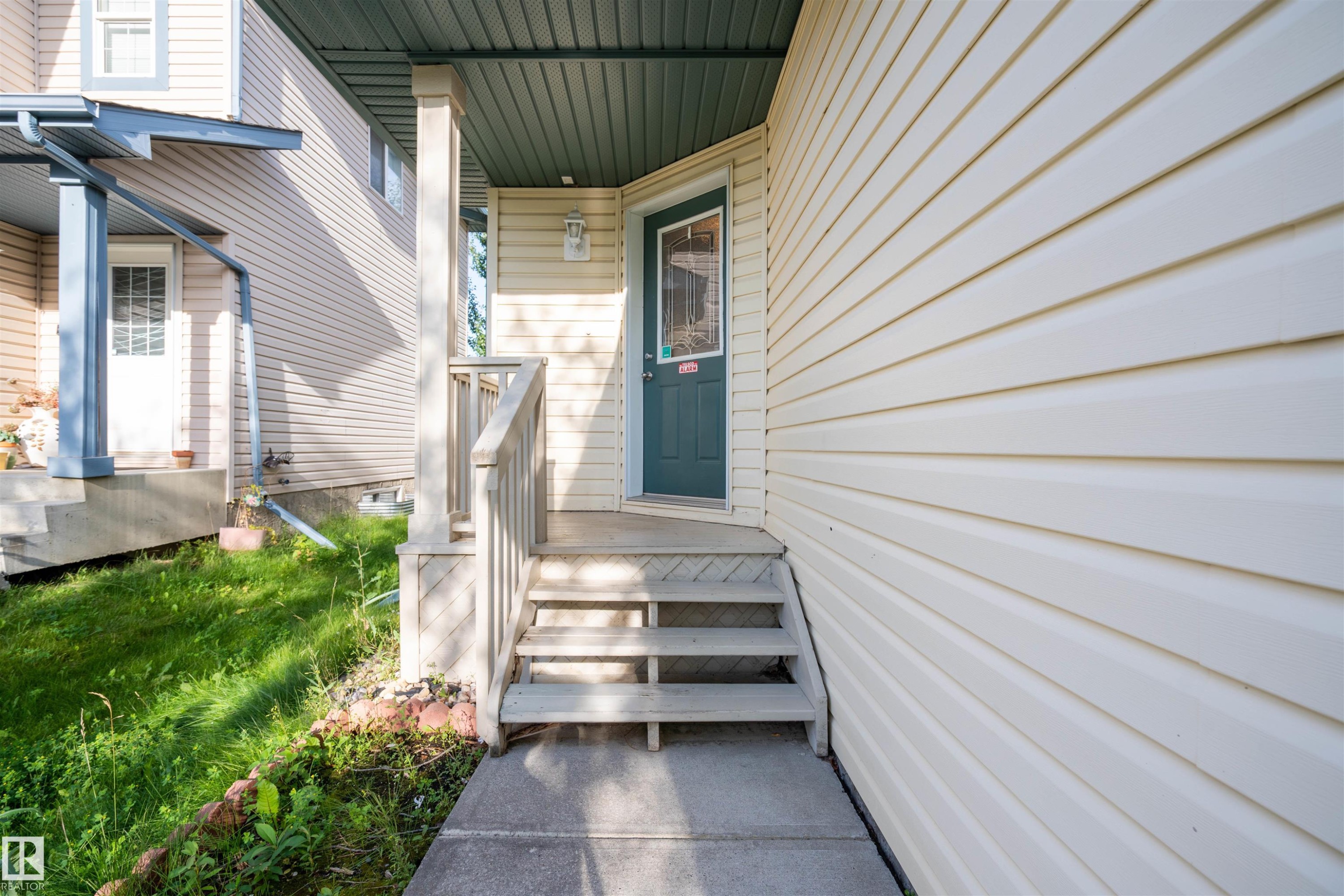 Inviting covered front porch with a dark green entry door, light-colored siding, concrete walkway, and wooden handrails - 1009 Macewan Close Sw, Edmonton, AB - Outdoor