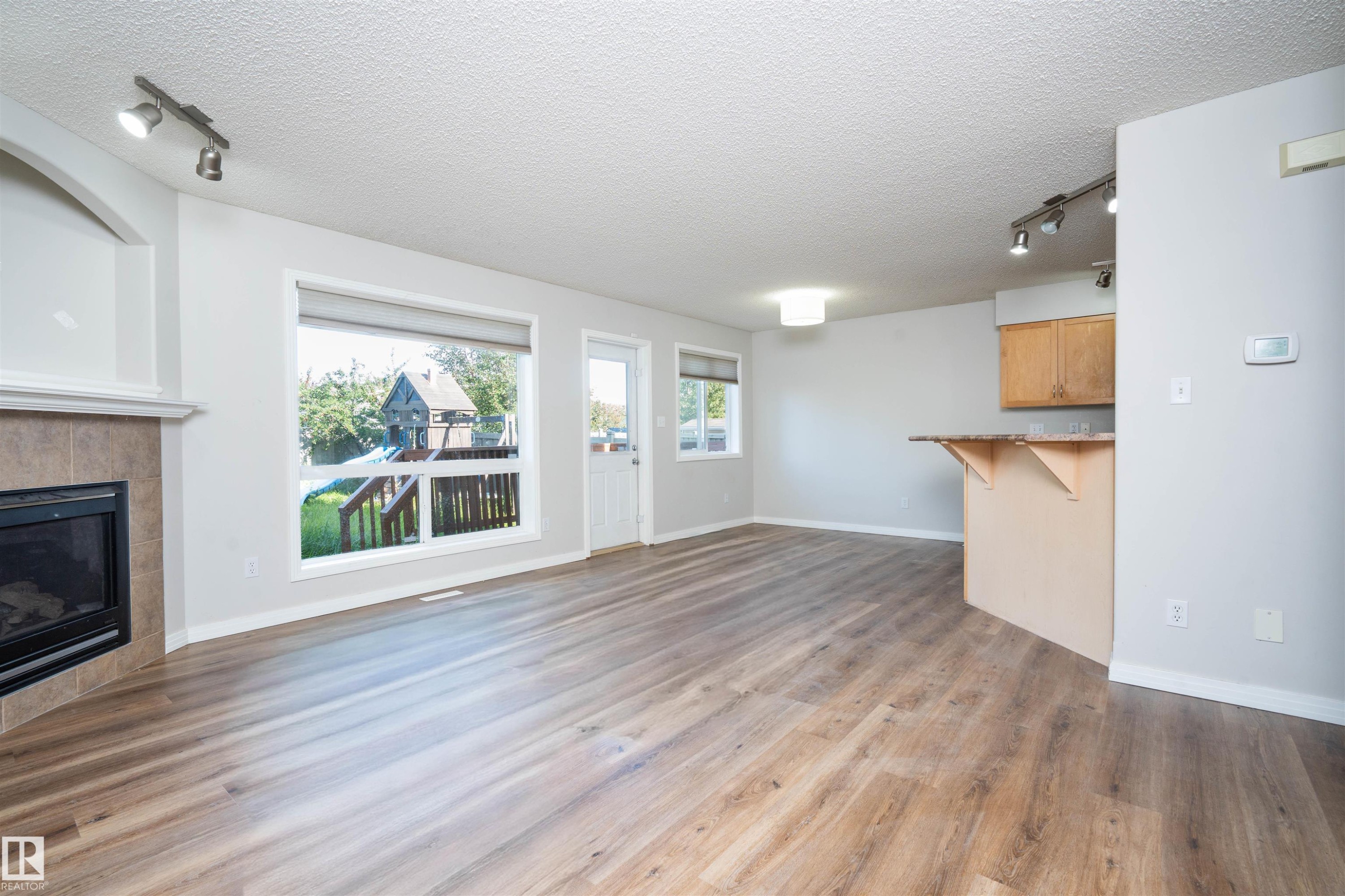 Spacious open concept room featuring wood-finish flooring, a tiled fireplace with mantel, multiple large windows, and track lighting - 1009 Macewan Close Sw, Edmonton, AB - Indoor Photo Showing Living Room With Fireplace