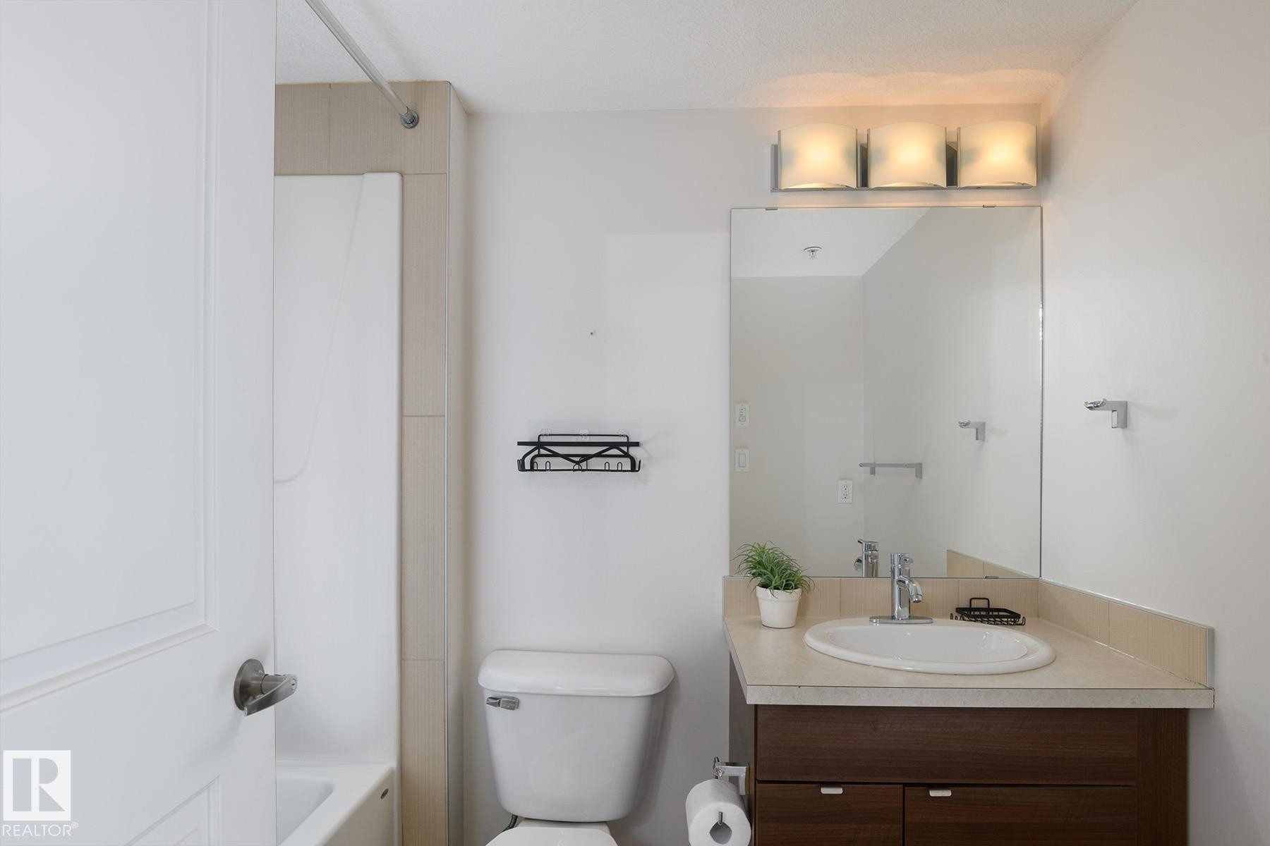 Bathroom featuring a wood-finish vanity with an integrated sink, a large wall-mounted mirror, and a three-light fixture - #110 9523 160 Av Nw, Edmonton, AB