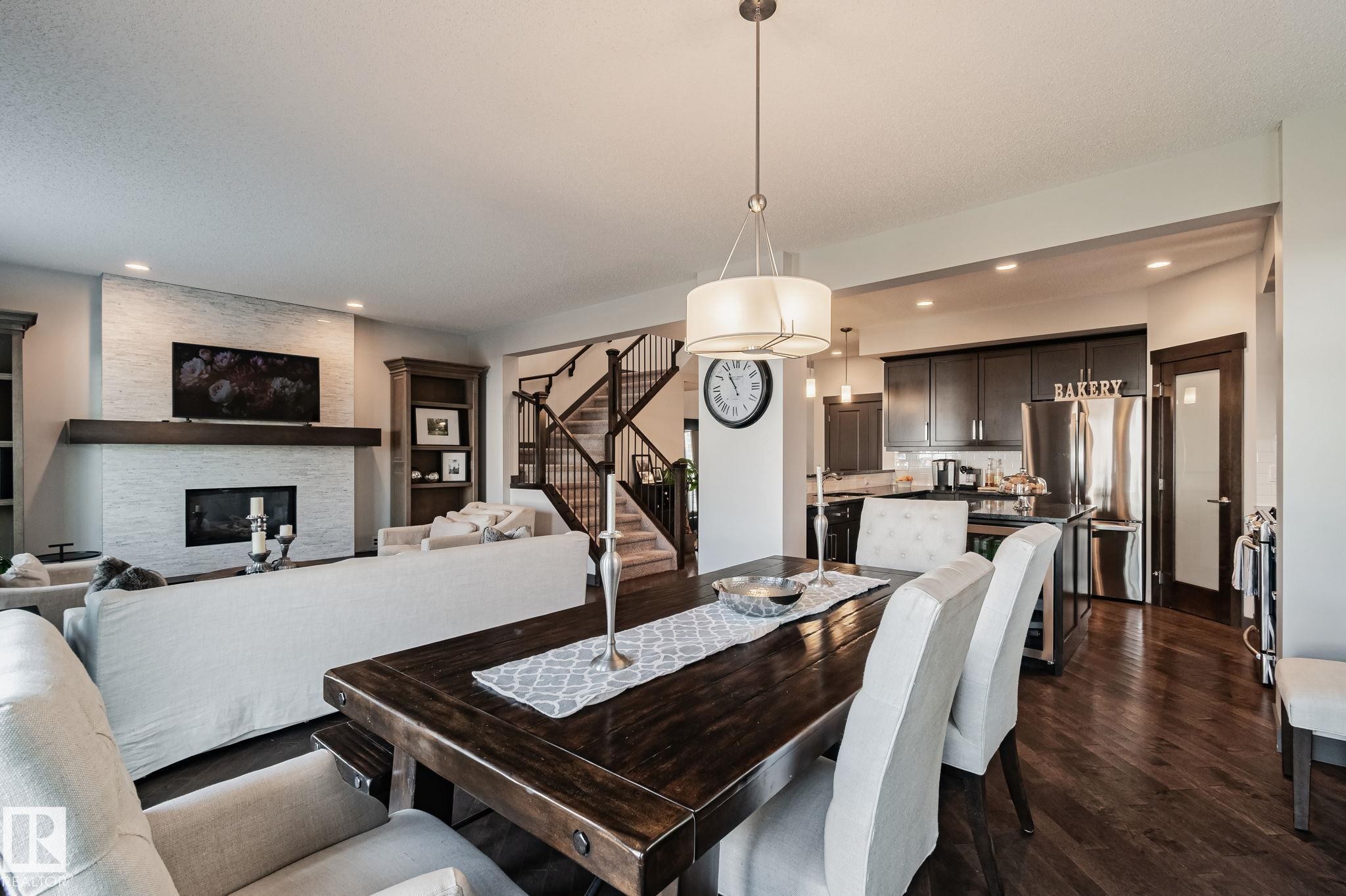Open-concept design featuring wood-finish flooring, a contemporary fireplace with a stone surround and wood mantle, a staircase with dark wood railings, and a kitchen with dark cabinetry and stainless steel appliances - 1337 Secord Landing Nw, Edmonton, AB - Indoor Photo Showing Other Room With Fireplace