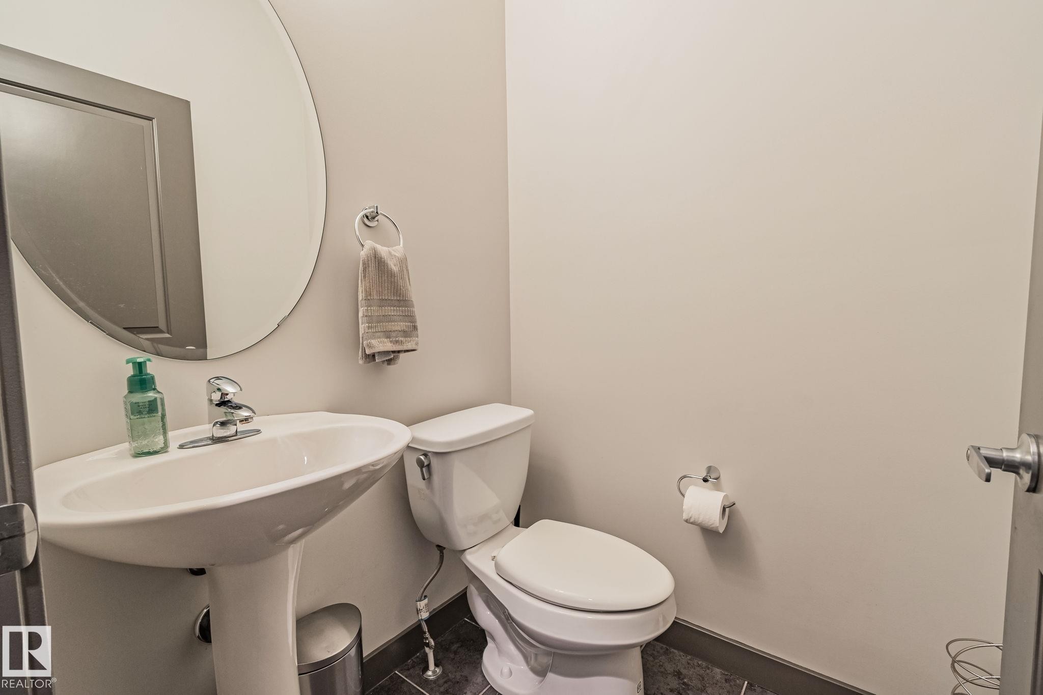 Pedestal sink with chrome faucet, circular wall mirror, and dark tile flooring - 1337 Secord Landing Nw, Edmonton, AB - Indoor Photo Showing Bathroom
