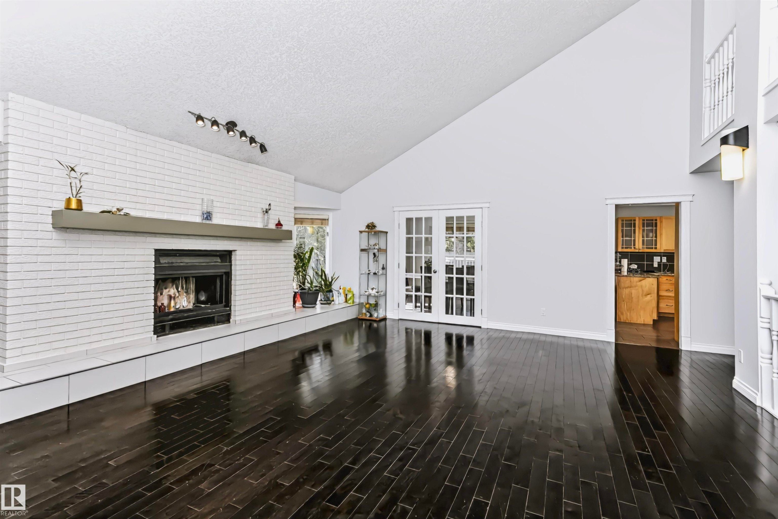 Spacious interior featuring a vaulted ceiling, dark wood-finish flooring, a white brick fireplace with a mantel, and double french doors - 6016 177 Street Nw, Edmonton, AB - Indoor With Fireplace