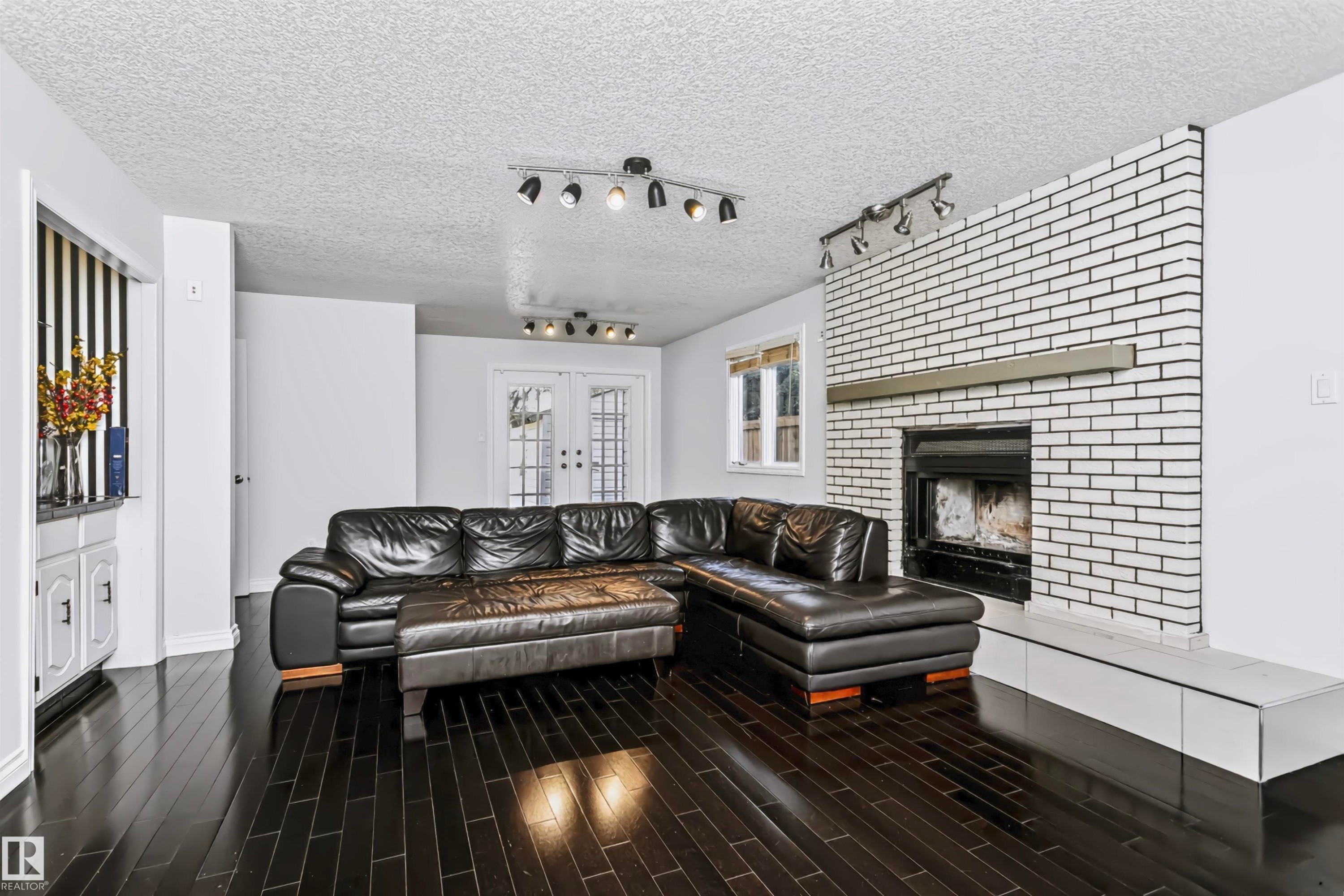 Dark wood-finish flooring, white painted brick fireplace with hearth, built-in white cabinetry, track lighting fixtures, and white French doors - 6016 177 Street Nw, Edmonton, AB - Indoor Photo Showing Living Room With Fireplace