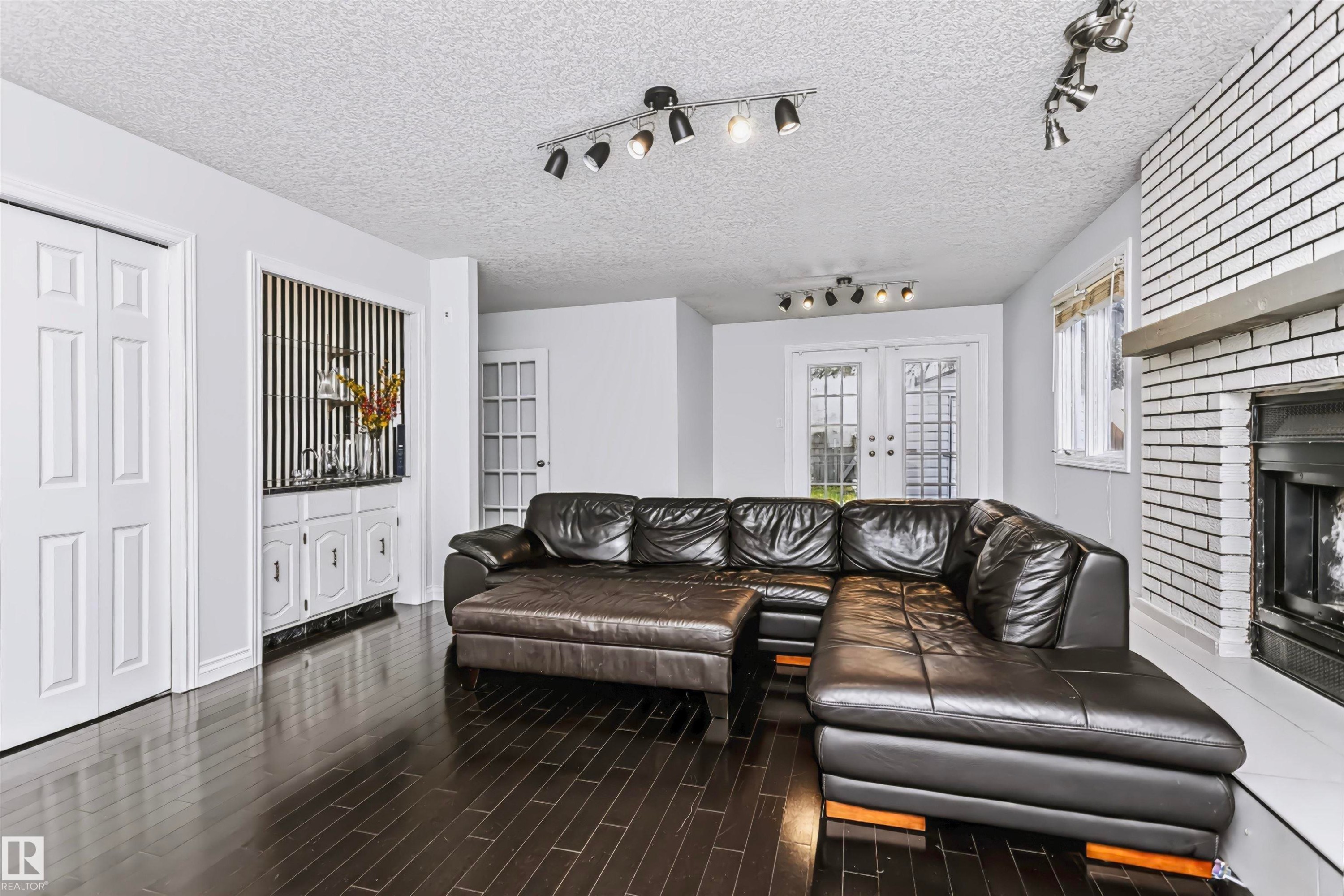 Dark wood-finish flooring, white painted brick fireplace with a mantel, built-in cabinetry, French doors, and track lighting fixtures - 6016 177 Street Nw, Edmonton, AB - Indoor Photo Showing Living Room With Fireplace