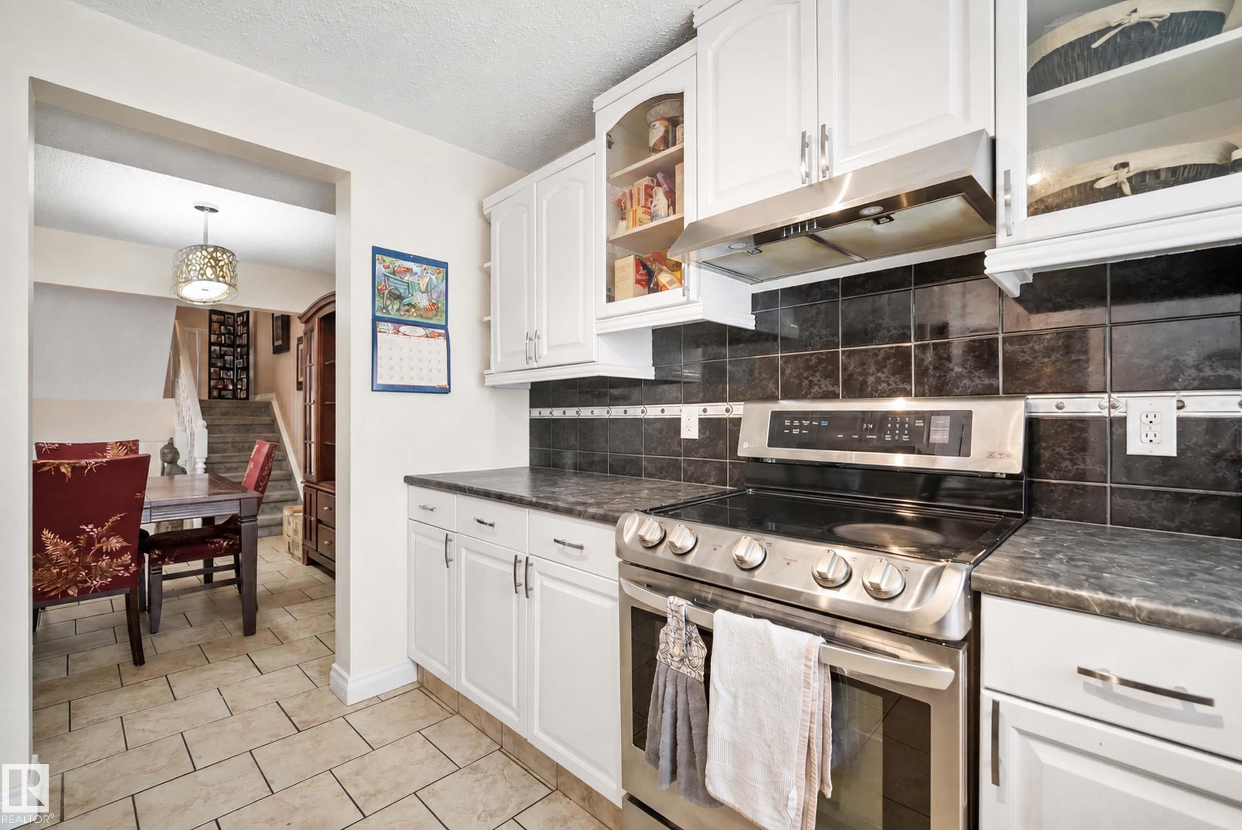 White cabinetry, black tile backsplash, stainless steel range with overhead vent, and dark countertops - #25 3811 85 St Nw, Edmonton, AB - Indoor Photo Showing Kitchen