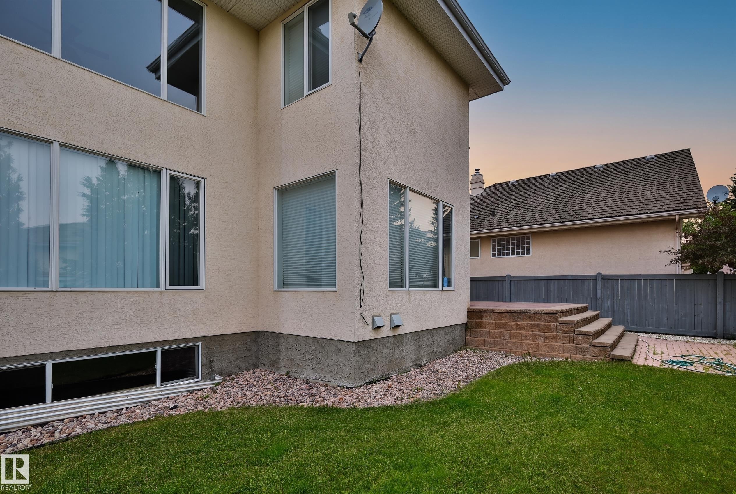 Stucco exterior with multiple windows, leading to a raised patio with brick-finish steps, a manicured lawn, and a privacy fence - 237 Darlington Crescent Nw, Edmonton, AB - Outdoor With Exterior