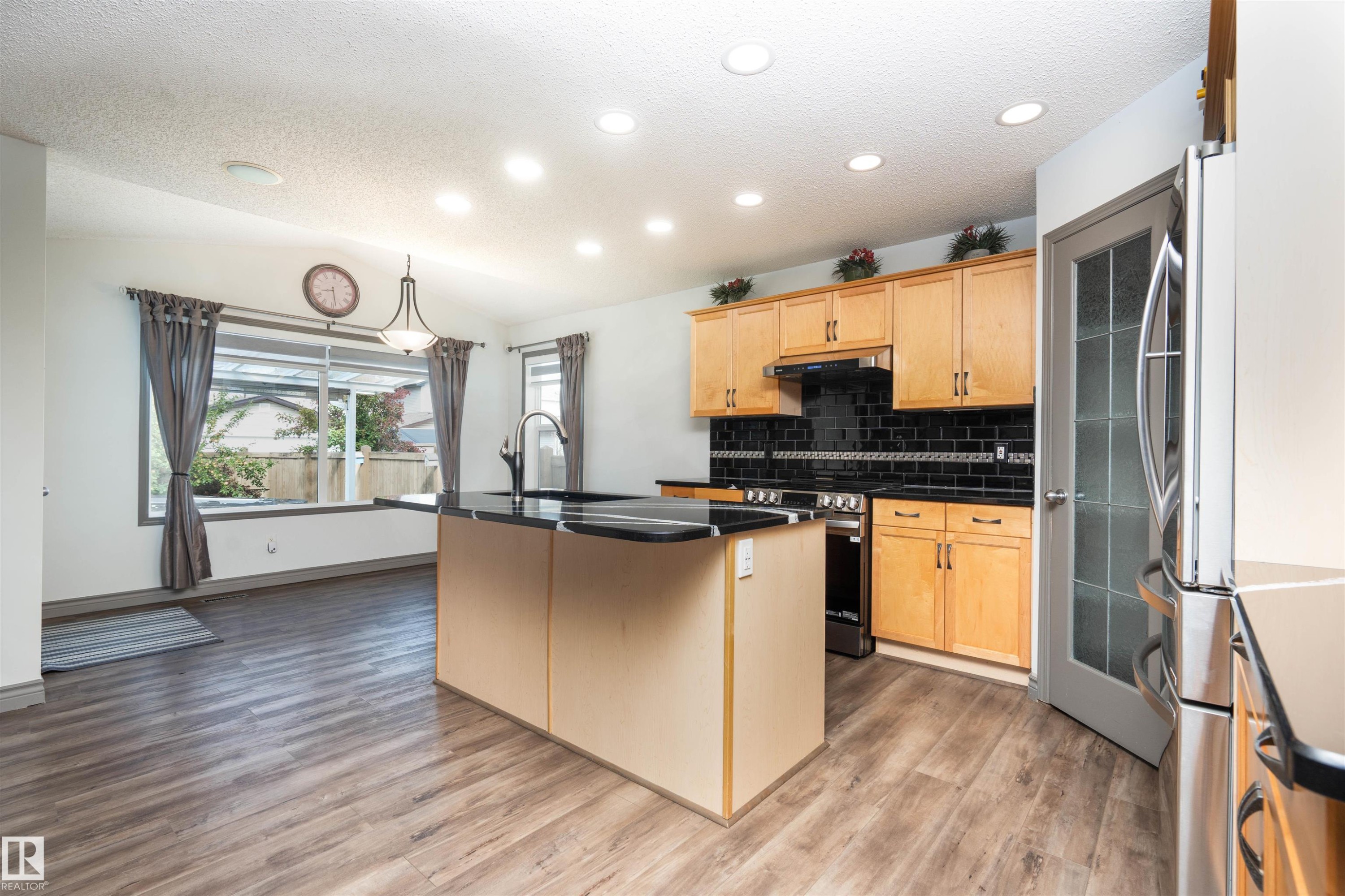 Kitchen featuring wood-finish flooring, recessed lighting, a central island with an integrated sink, black countertops, and light wood cabinetry with a dark tile backsplash - 16820 57 Street Nw, Edmonton, AB - Indoor Photo Showing Kitchen