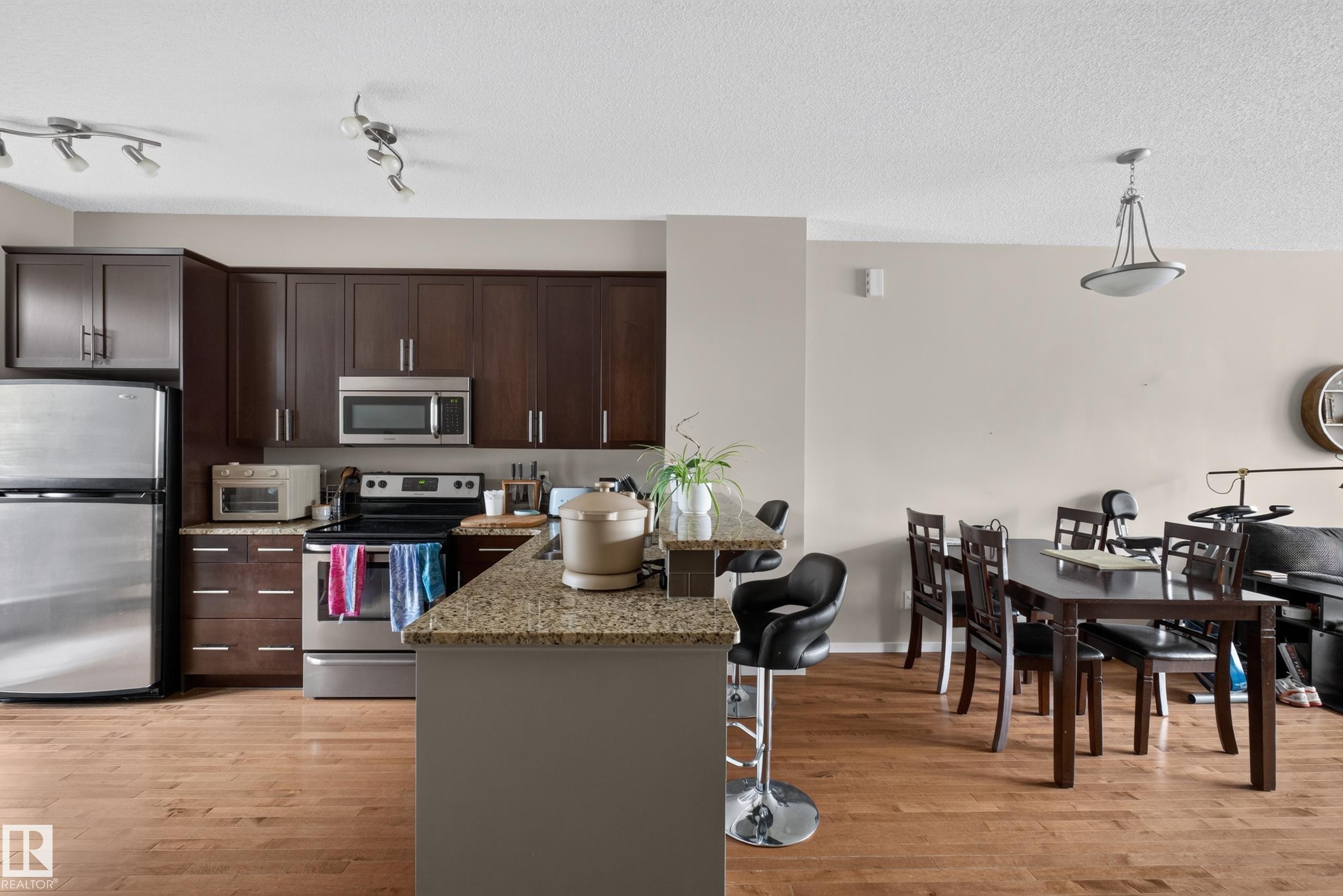 Open-concept kitchen and dining area featuring wood-finish flooring, dark wood cabinetry, stainless steel appliances, and track lighting - #53 603 Watt Bv Sw, Edmonton, AB - Indoor Photo Showing Kitchen