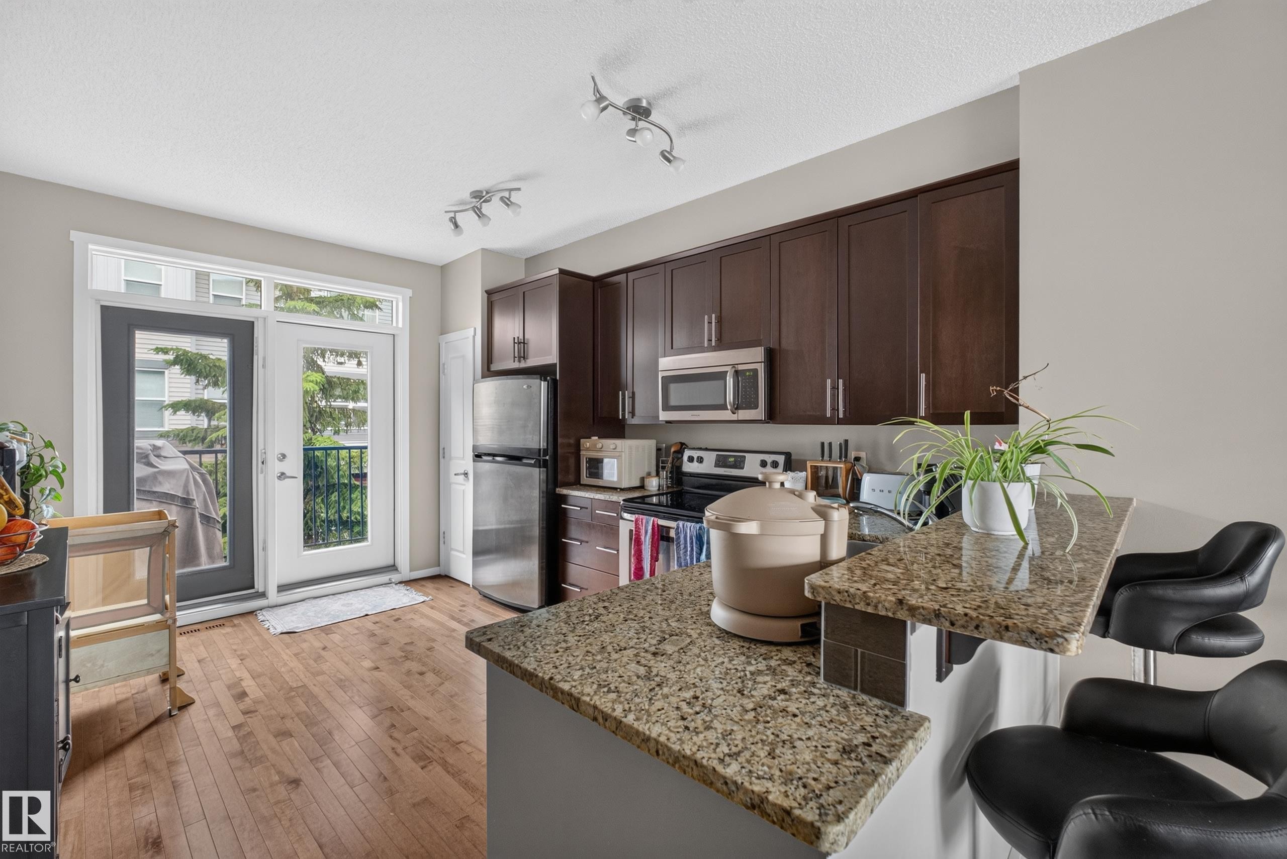 Kitchen featuring wood-finish flooring, dark wood cabinetry, stainless steel appliances, and granite countertops with a breakfast bar - #53 603 Watt Bv Sw, Edmonton, AB - Indoor Photo Showing Kitchen