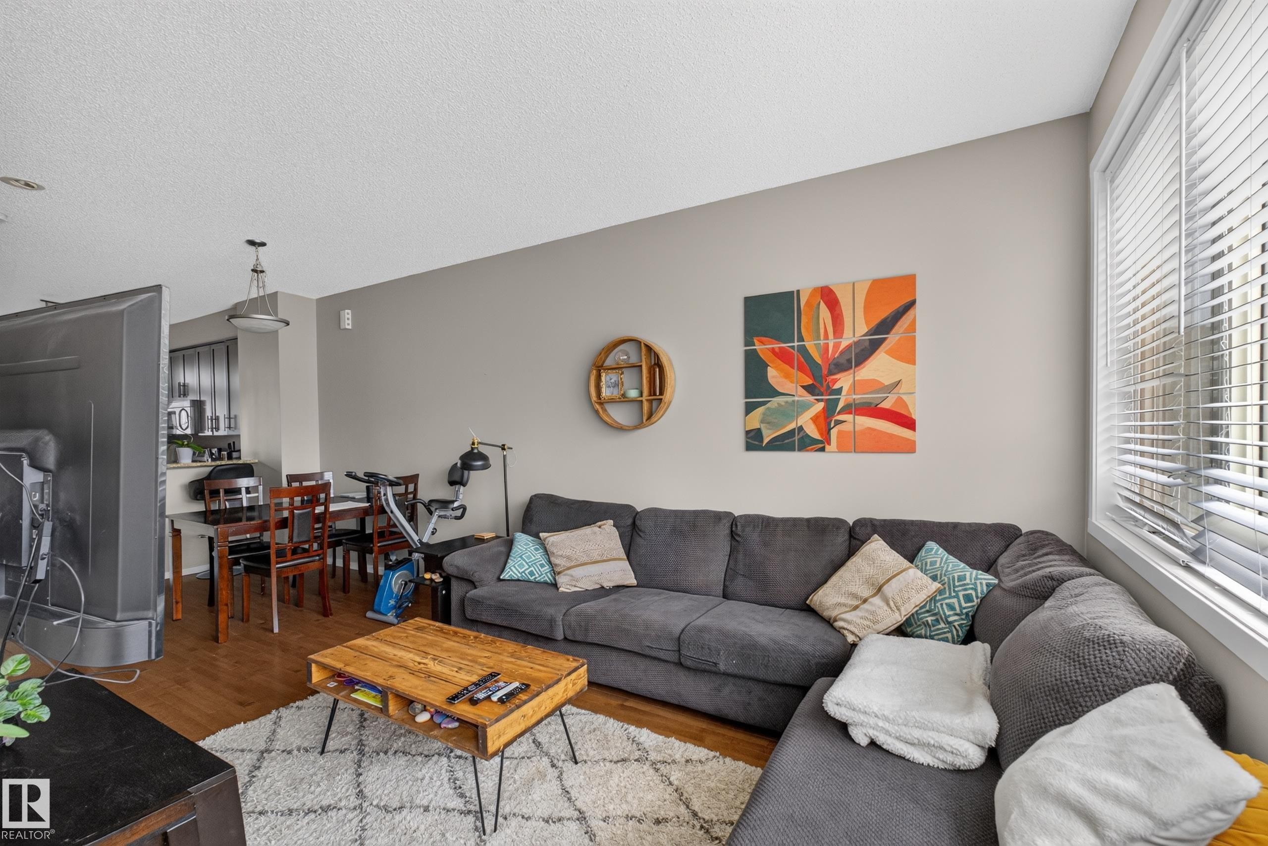 Living area with wood-finish flooring, light-toned walls, and a large window with horizontal blinds - #53 603 Watt Bv Sw, Edmonton, AB - Indoor Photo Showing Living Room