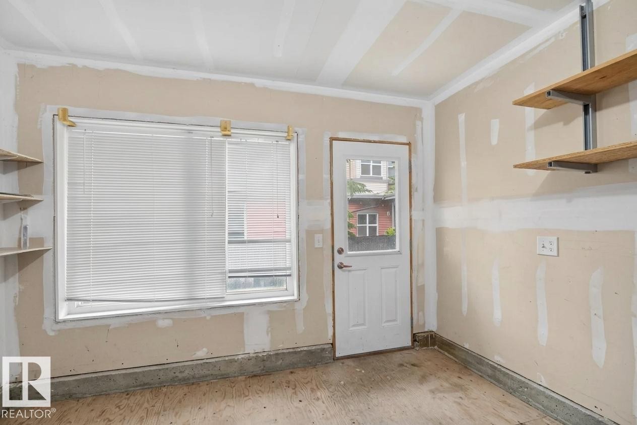 Versatile room featuring exposed drywall, a window with blinds, a white door with a glass pane, plywood flooring, and built-in shelving - #53 603 Watt Bv Sw, Edmonton, AB - Indoor Photo Showing Other Room