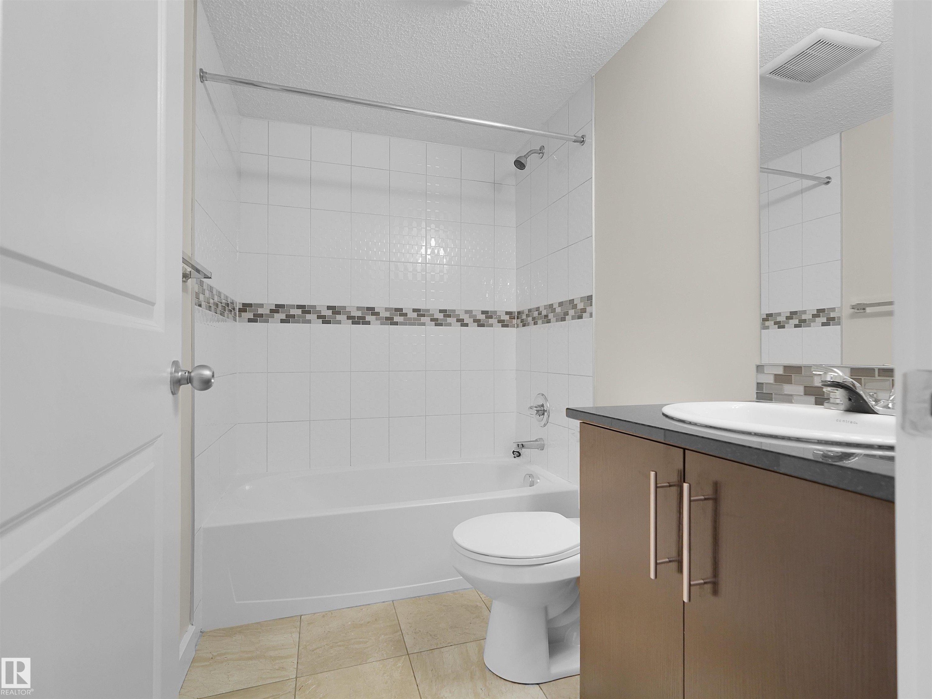White tile surround shower-tub combination, mosaic tile accent, wood-finish vanity with countertop, oval undermount sink, and tile flooring - #220 5804 Mullen Pl Nw, Edmonton, AB