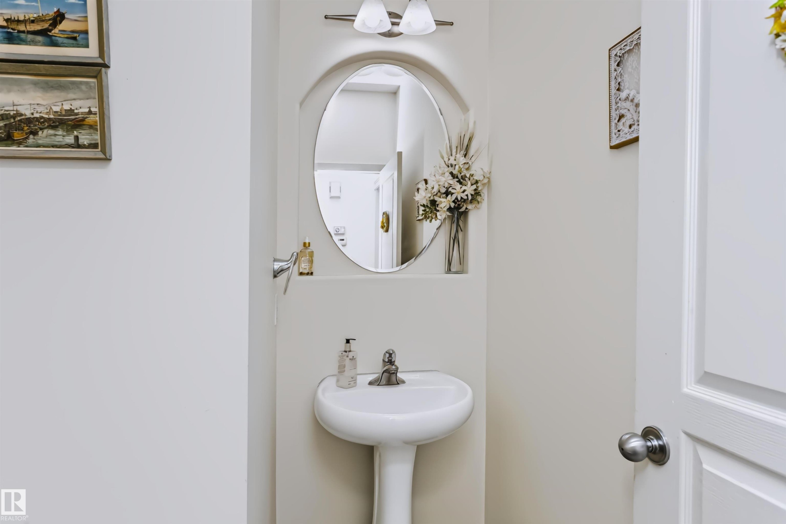 Pedestal sink with a polished chrome faucet, oval mirror, and wall-mounted vanity lighting - 1720 Robertson Place Sw, Edmonton, AB - Indoor Photo Showing Bathroom