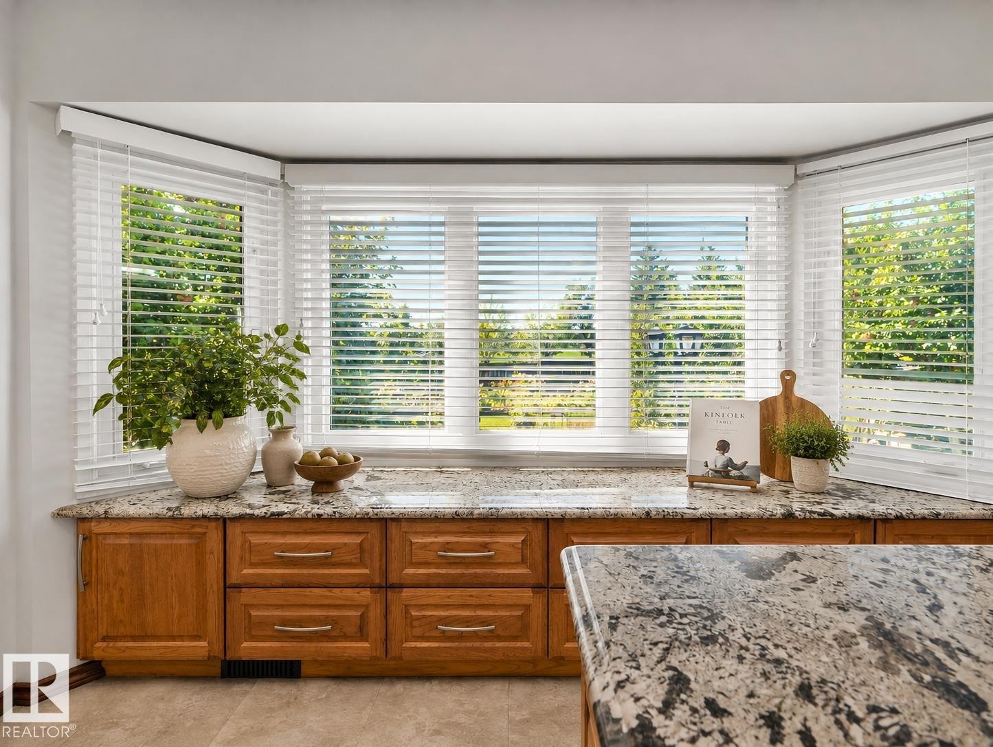 Bright bay window featuring multiple panes of plantation shutters, polished stone countertop, and warm wood-finish cabinetry with integrated drawer storage - 7357 18 Avenue Nw, Edmonton, AB - Indoor