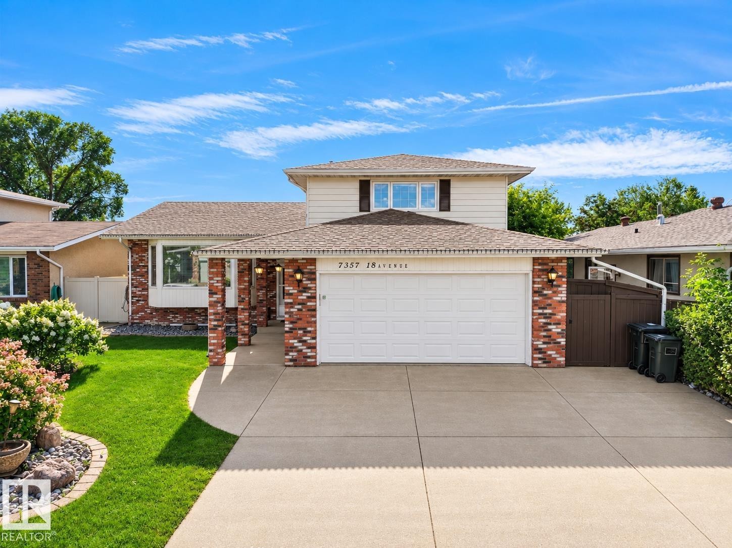 Two-story brick exterior with white garage doors, manicured front lawn, mature landscaping, concrete driveway, and second-story dormer windows - 7357 18 Avenue Nw, Edmonton, AB - Outdoor