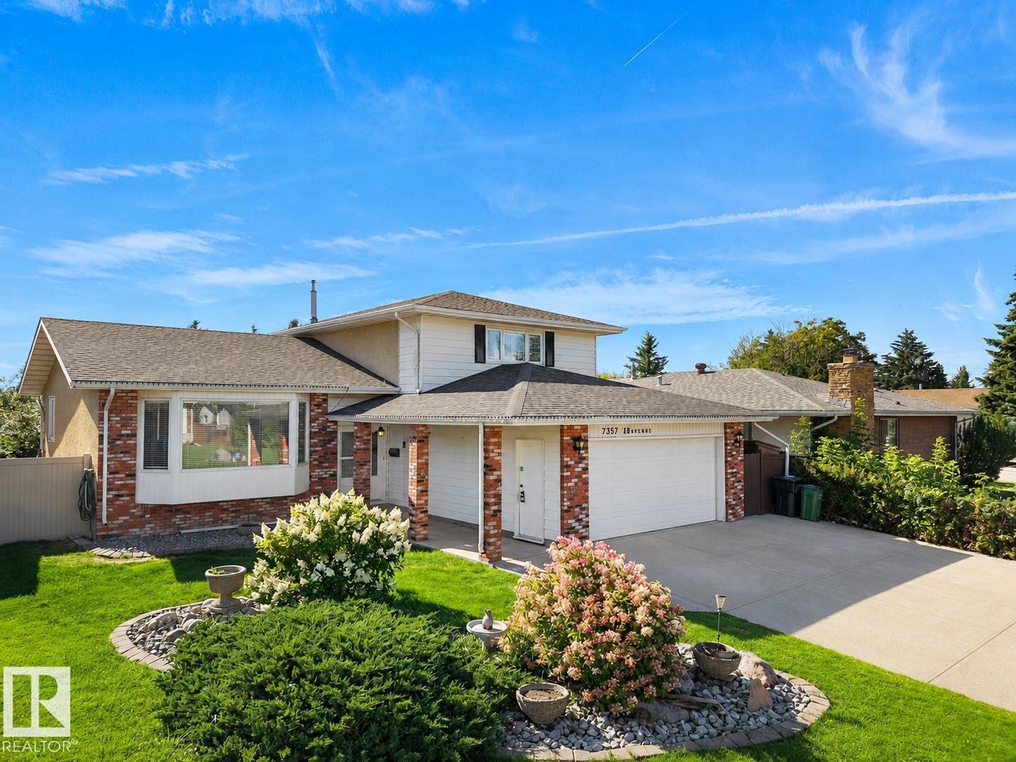 Welcoming two-story residence featuring a brick and stone exterior with multiple windows, a two-car attached garage, and a professionally landscaped front yard with mature plantings, decorative stone accents, and a manicured lawn - 7357 18 Avenue Nw, Edmonton, AB - Outdoor With Facade