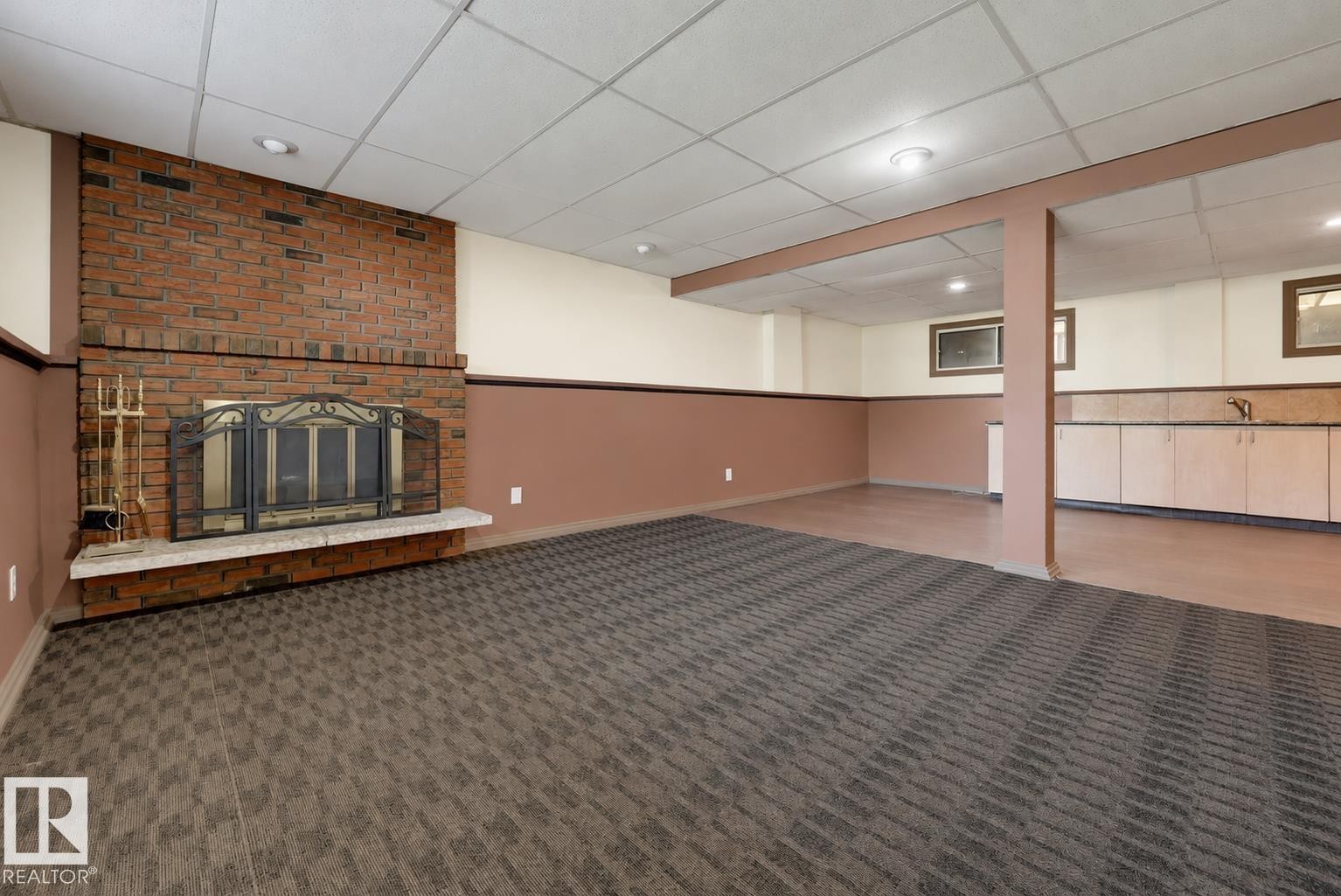 Spacious basement recreation room featuring exposed brick fireplace with decorative surround, neutral taupe wall paneling, patterned carpet flooring, recessed ceiling lighting, and abundant square footage ideal for entertaining or flexible living space - 6768 39 Avenue Nw, Edmonton, AB - Indoor With Fireplace