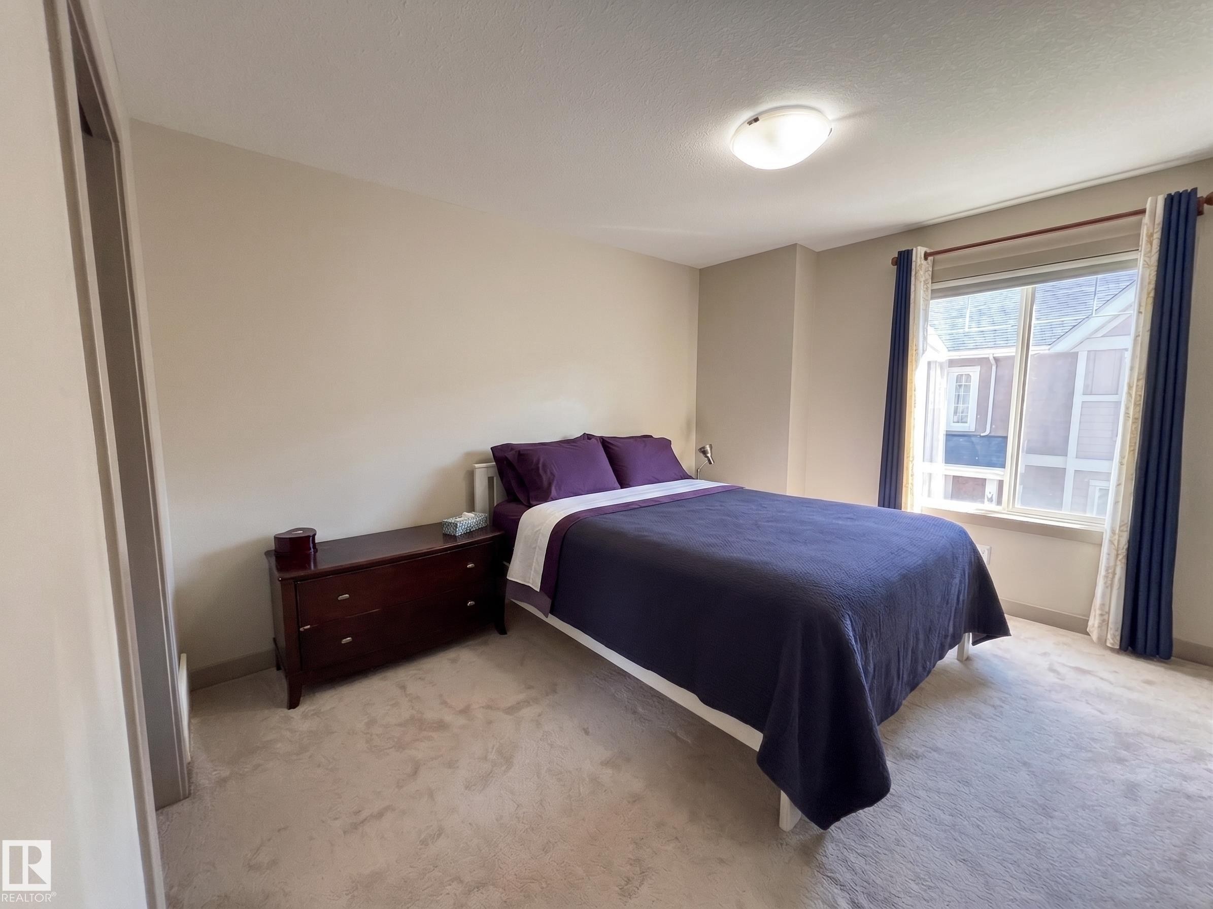 Spacious secondary bedroom featuring neutral wall finishes, carpeted flooring, large windows with blue drapery, and dark wood bedside furnishings - #29 1150 Windermere Wy Sw, Edmonton, AB