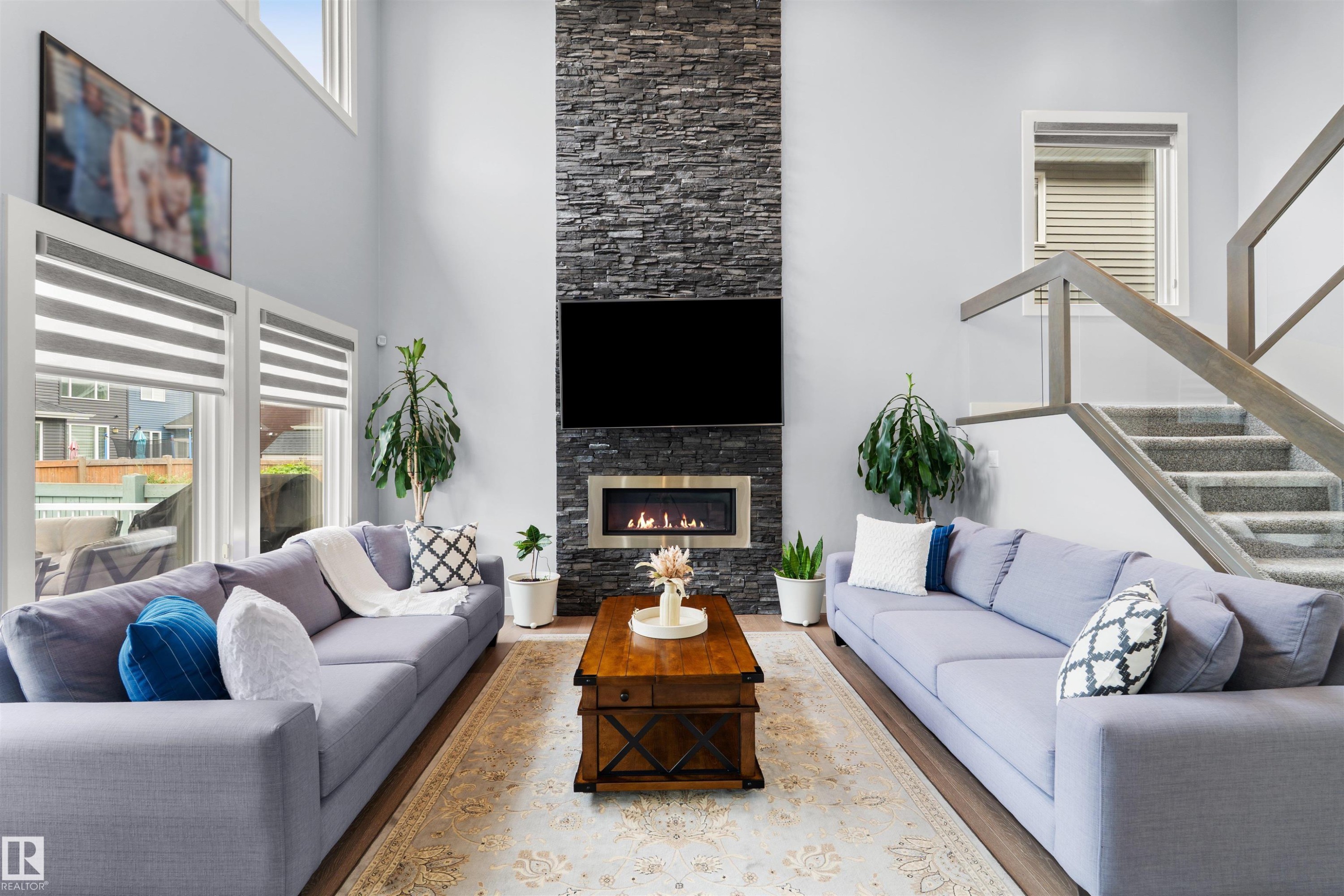 Vaulted ceiling, stone-clad fireplace with built-in insert, large windows, wood-finish flooring, and a staircase with glass panel railing - 16327 17 Avenue Sw, Edmonton, AB - Indoor Photo Showing Living Room With Fireplace