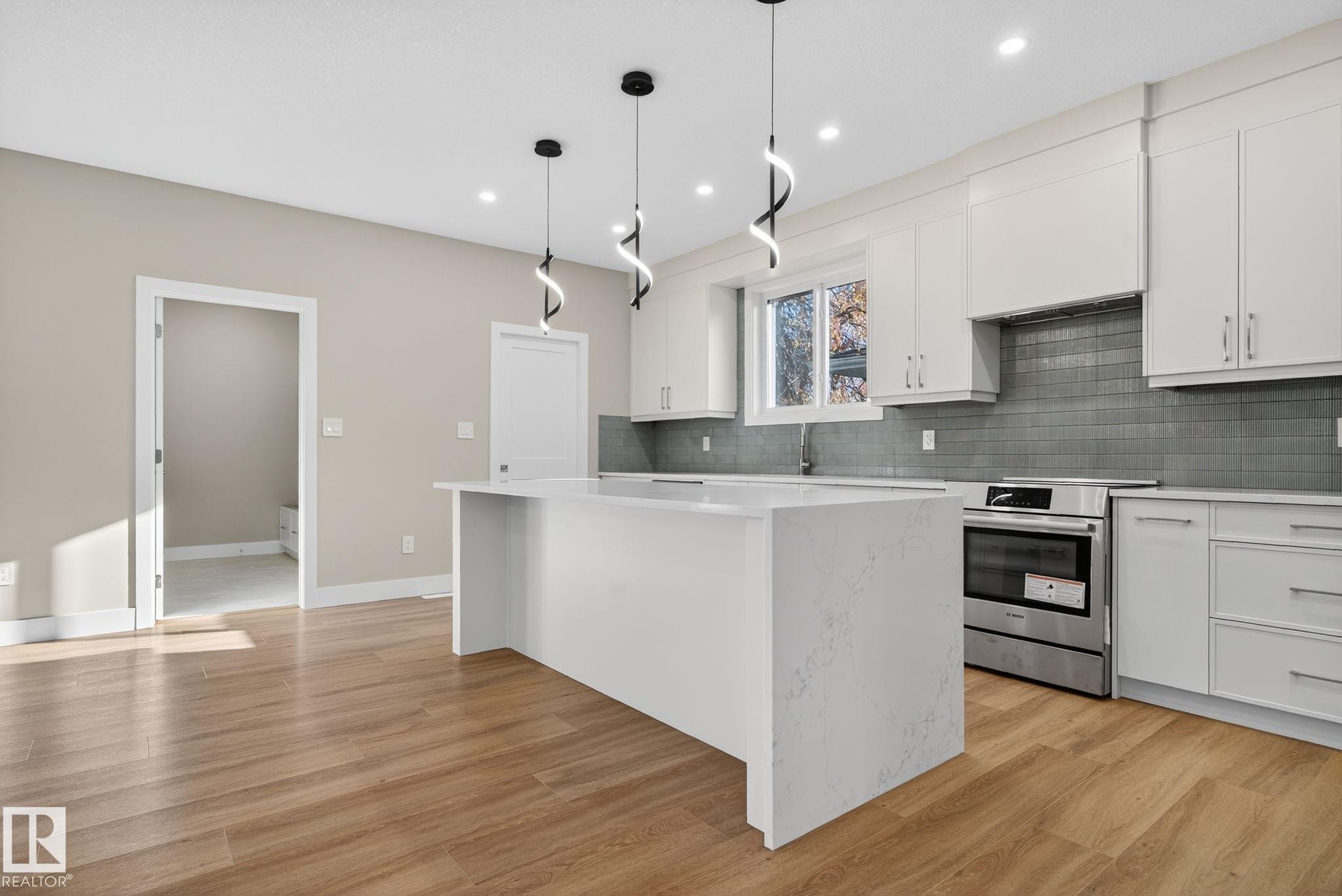 Modern kitchen featuring white cabinetry, a large center island with a light-toned countertop, a stainless steel range, and wood-finish flooring - 9736 73 Avenue Nw, Edmonton, AB