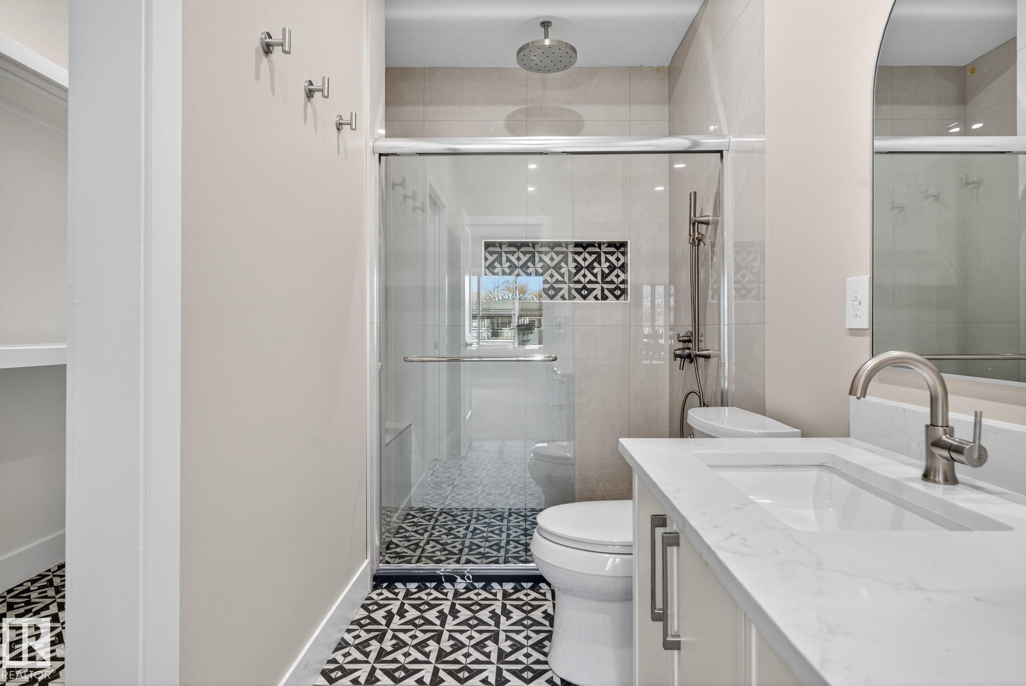 Patterned floor tiling, glass-enclosed shower, rain shower head, built-in shower niche with decorative tiling, and a vanity with an integrated sink and brushed nickel faucet - 9736 73 Avenue Nw, Edmonton, AB