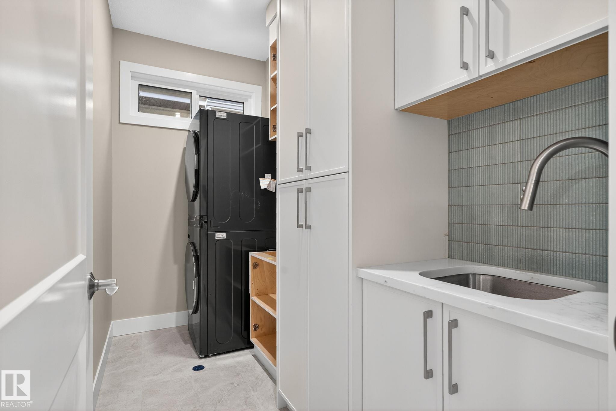 Laundry area featuring stacked appliances, white cabinetry with silver hardware, a utility sink with a gooseneck faucet, and a tiled backsplash - 9736 73 Avenue Nw, Edmonton, AB