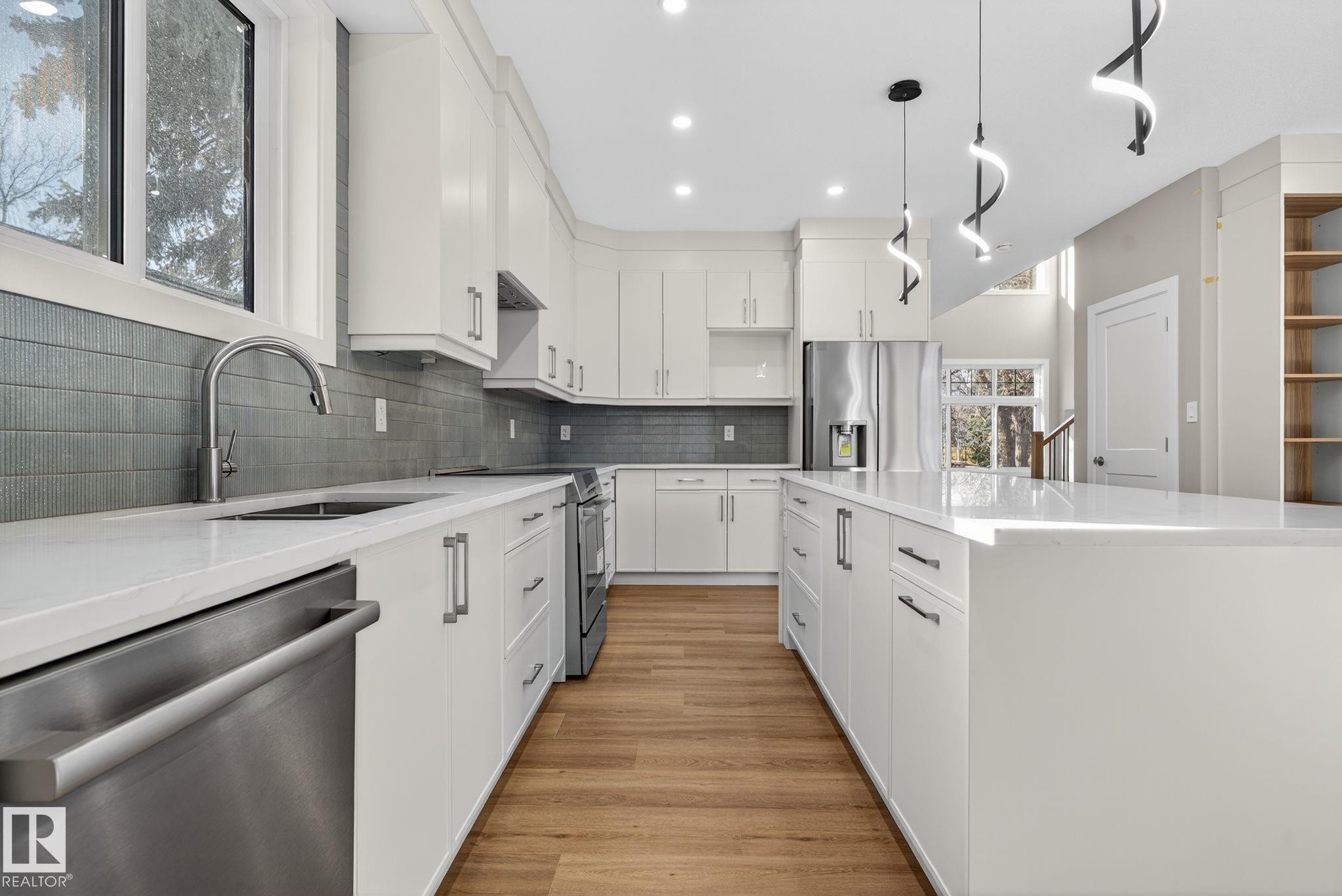 Modern kitchen featuring white cabinetry, light-toned wood-finish flooring, a kitchen island, stainless steel appliances, and sleek pendant lighting - 9736 73 Avenue Nw, Edmonton, AB