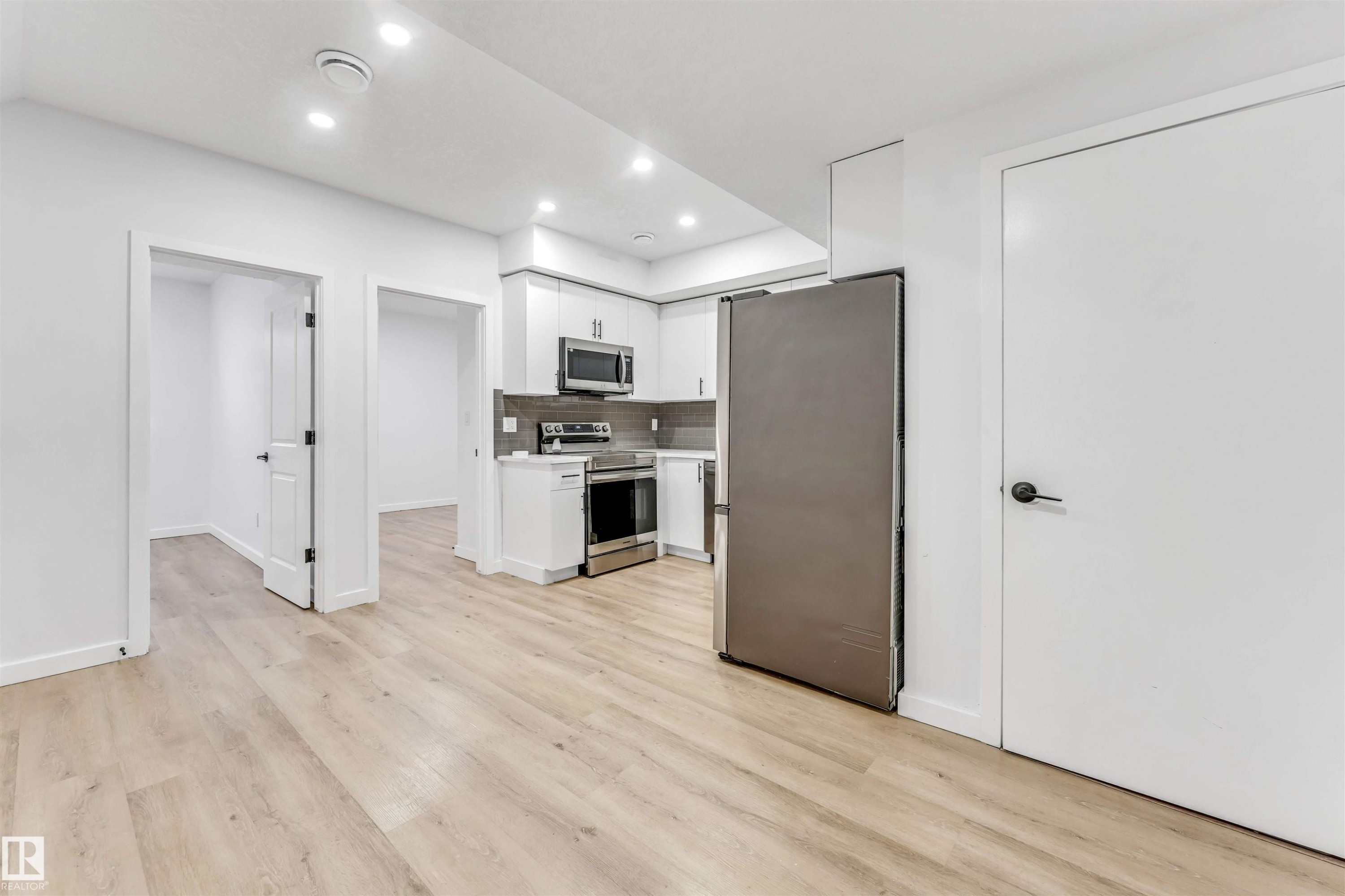 Basement: Wood-finish flooring throughout, recessed ceiling lighting, stainless steel appliances, white cabinetry, and a dark tile backsplash - 3236 Chernowski Way Sw, Edmonton, AB - Indoor Photo Showing Kitchen
