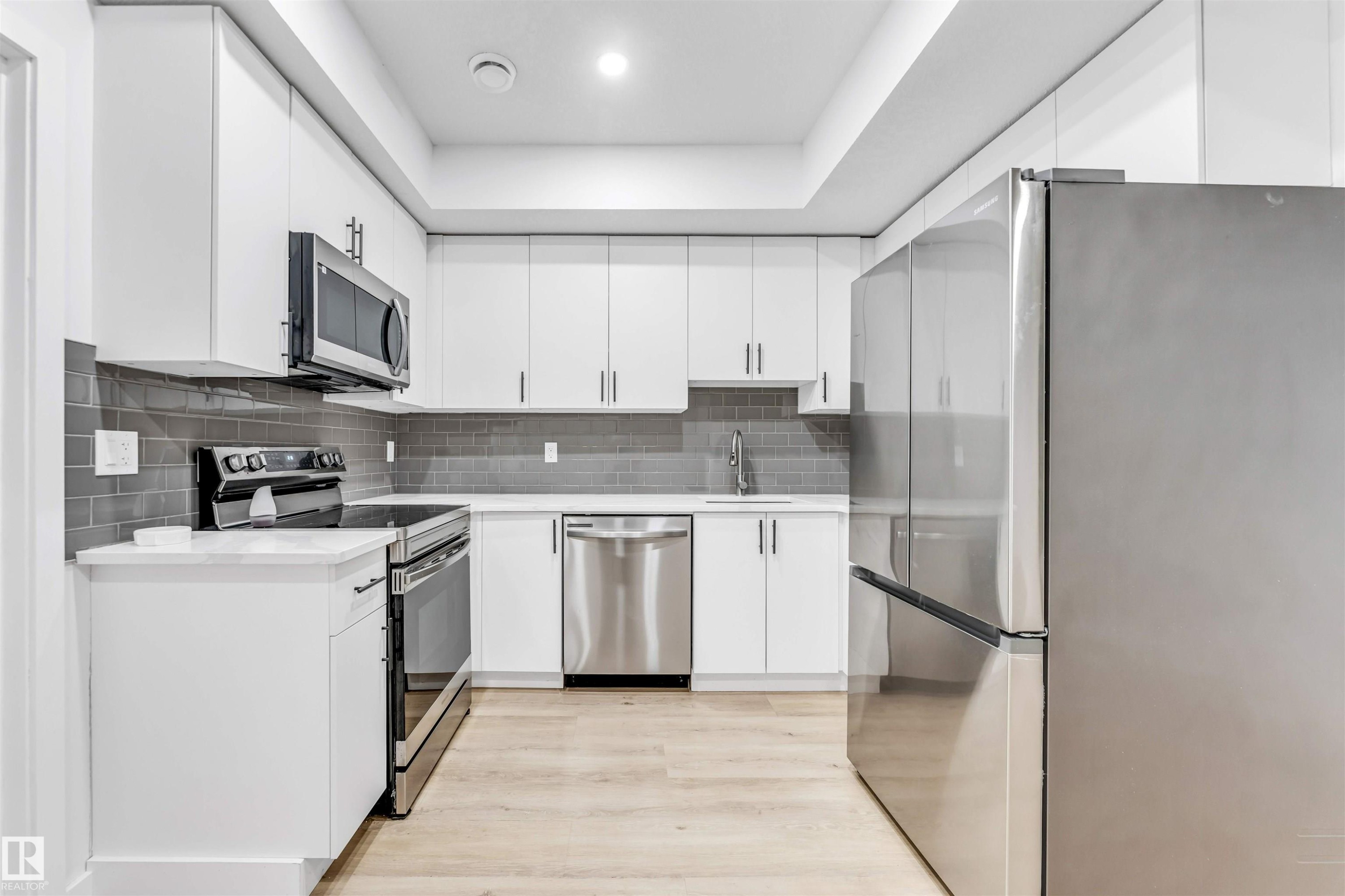 U-shaped kitchen with white cabinetry, light wood-finish flooring, stainless steel appliances, white countertops, and a gray subway tile backsplash - 3236 Chernowski Way Sw, Edmonton, AB - Indoor Photo Showing Kitchen With Upgraded Kitchen