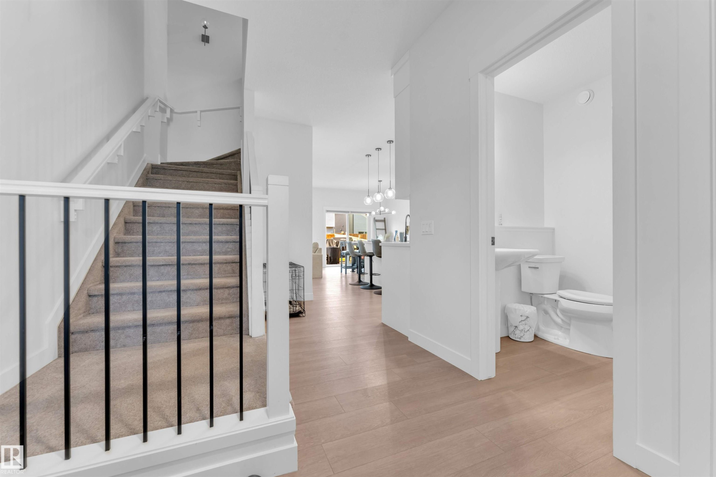 Wood-finish flooring, staircase with carpeted treads and black metal spindles, white painted handrail, and a half-bath with a white toilet and pedestal sink - 3236 Chernowski Way Sw, Edmonton, AB - Indoor Photo Showing Other Room