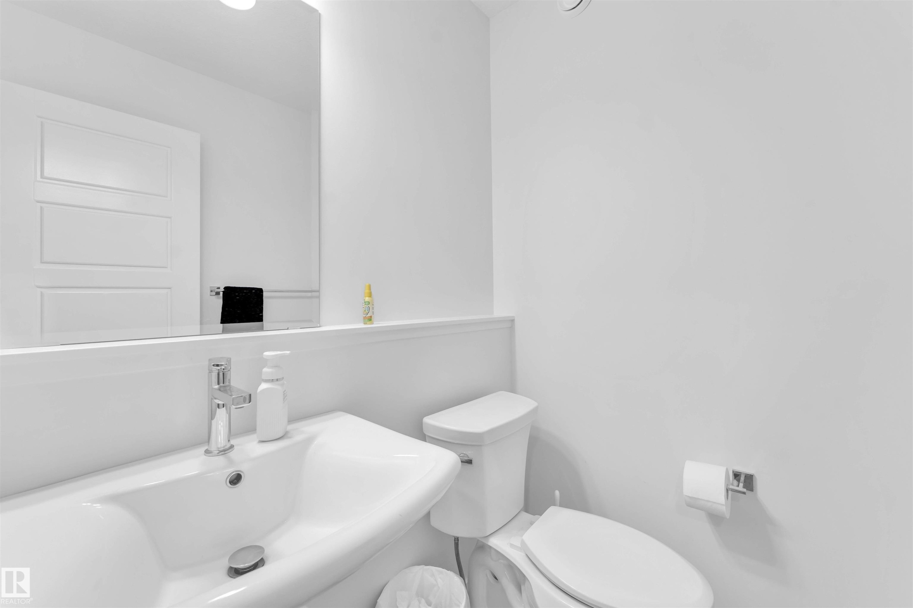 Pedestal sink with chrome faucet, wall-mounted mirror, white paneled door, and a wall-mounted toilet paper holder - 3236 Chernowski Way Sw, Edmonton, AB - Indoor Photo Showing Bathroom
