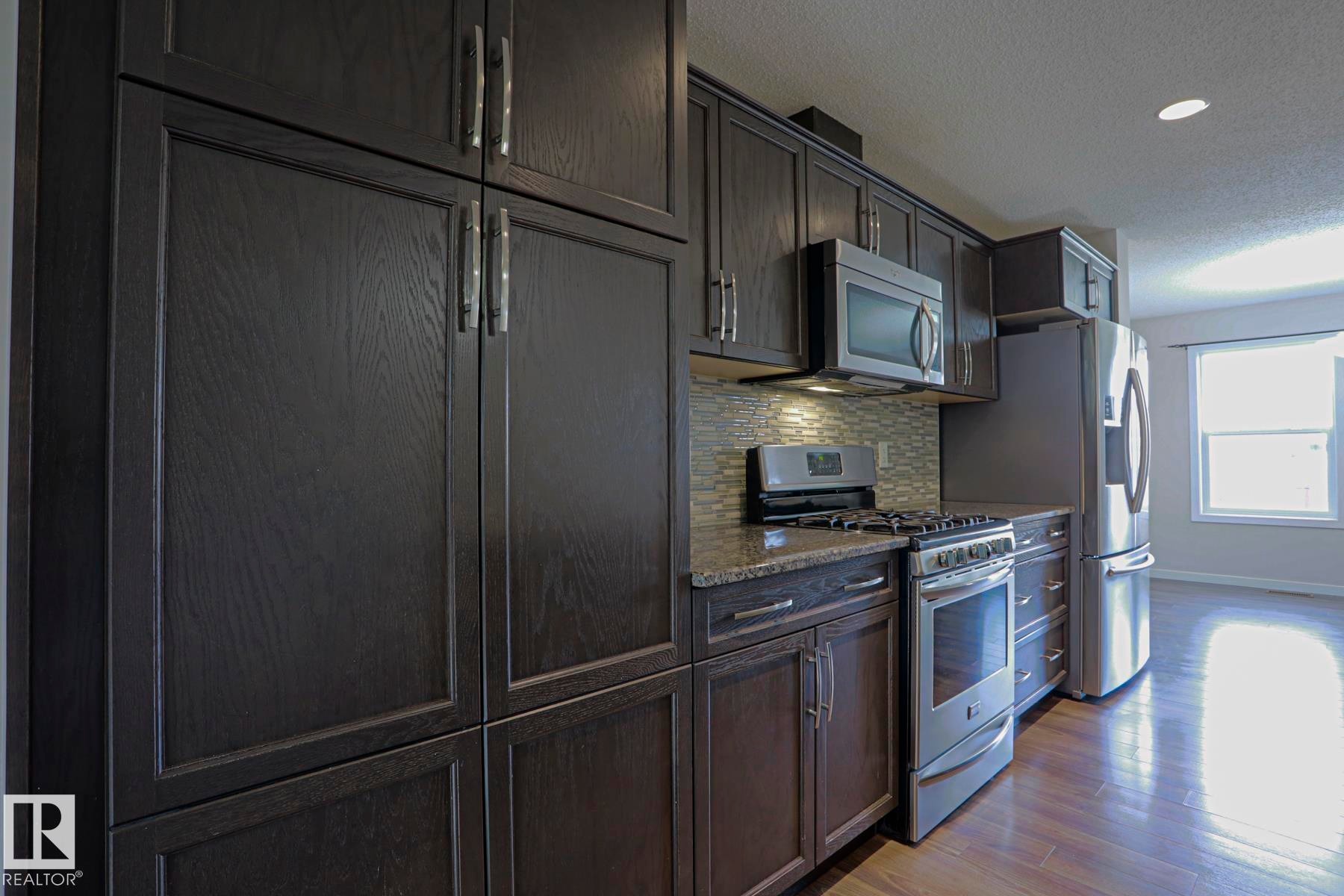 Kitchen featuring dark wood-finish cabinetry, stainless steel appliances, granite countertops, and a tiled backsplash - 308 Watt Boulevard Sw, Edmonton, AB - Indoor Photo Showing Kitchen