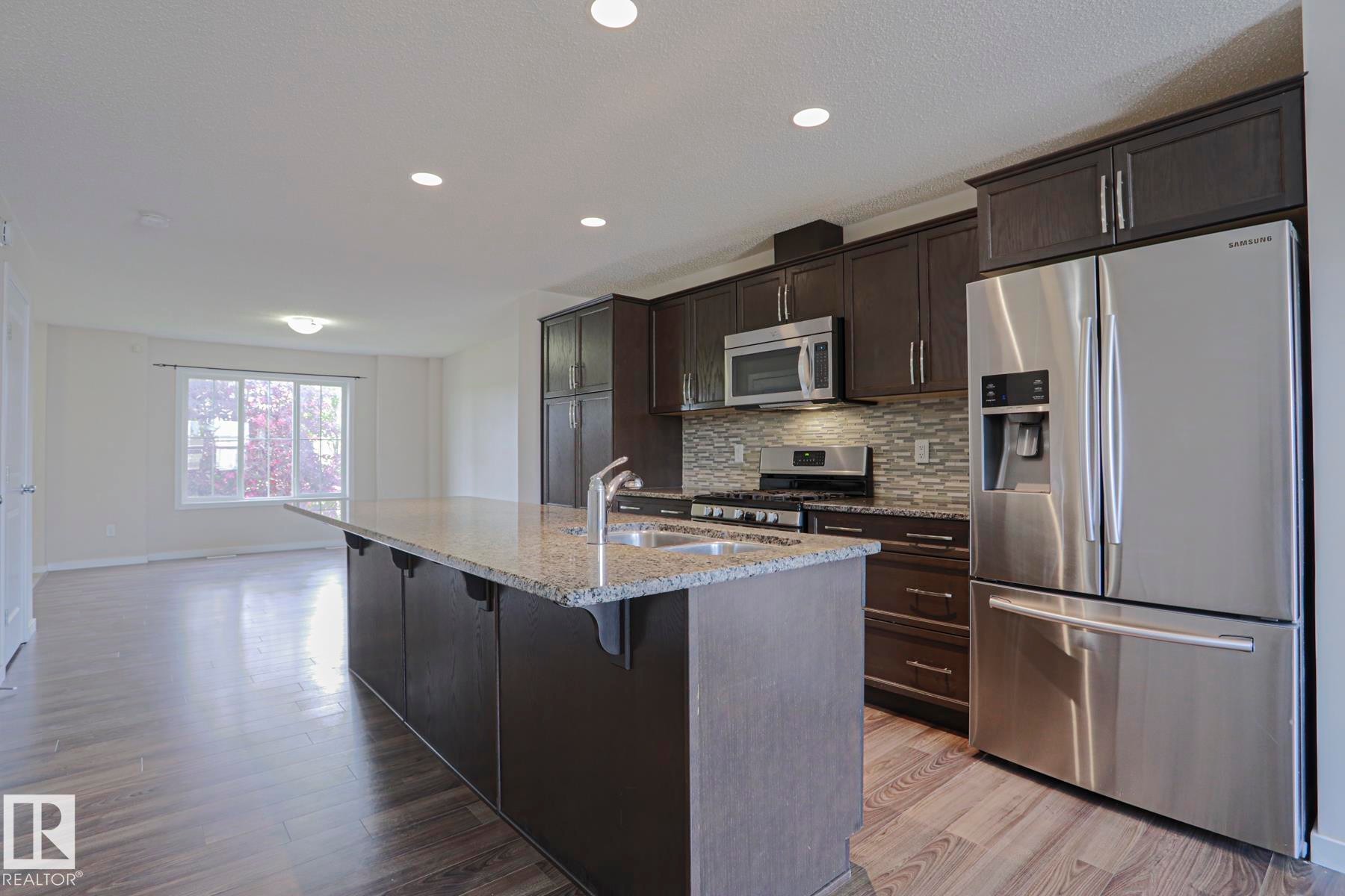 Kitchen featuring dark wood-finish cabinetry, stainless steel appliances, a tiled backsplash, and an island with a light-toned countertop - 308 Watt Boulevard Sw, Edmonton, AB - Indoor Photo Showing Kitchen With Upgraded Kitchen